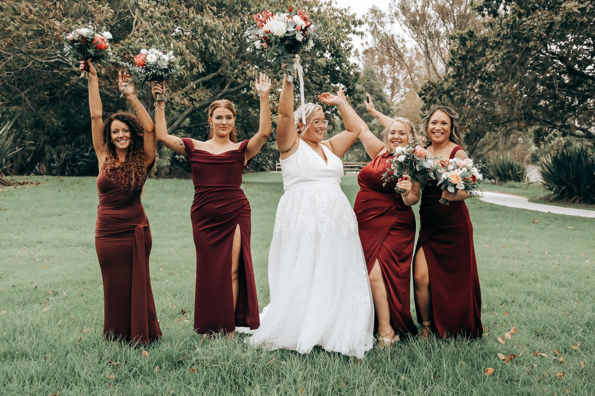 A bride and four bridesmaids holding bouquets, celebrating outdoors on a grassy area surrounded by trees.