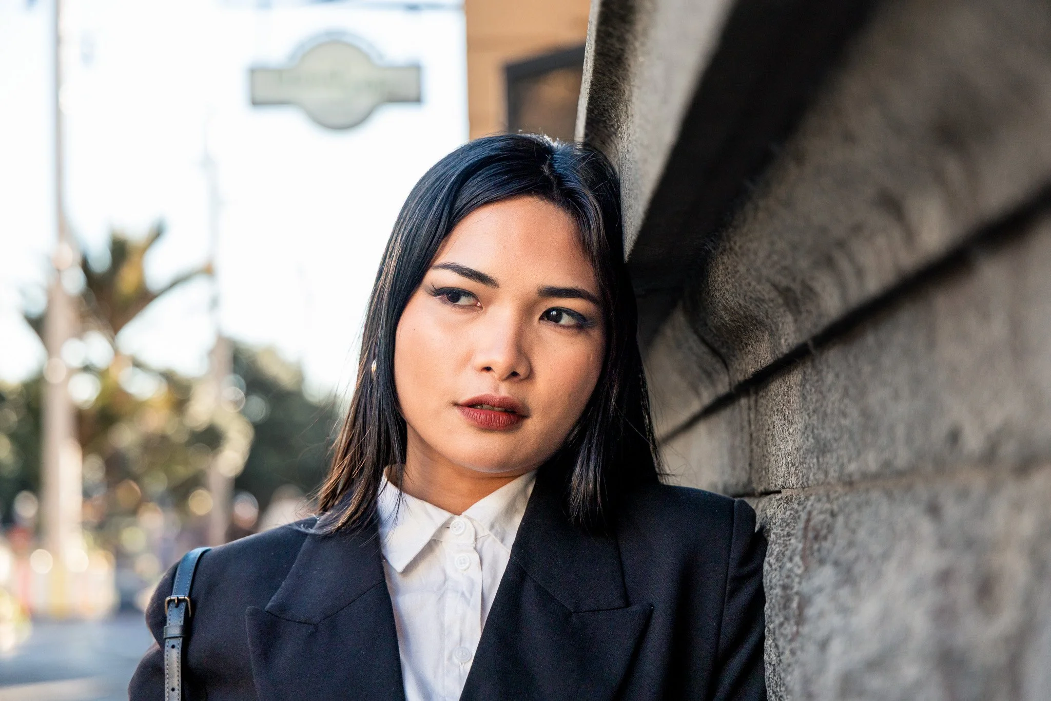 A young woman with black hair, wearing a black blazer over a white collared shirt, is leaning against a concrete wall outdoors. She has a backpack on her shoulder and is looking slightly to the side. There is a blurred background with trees and a bui