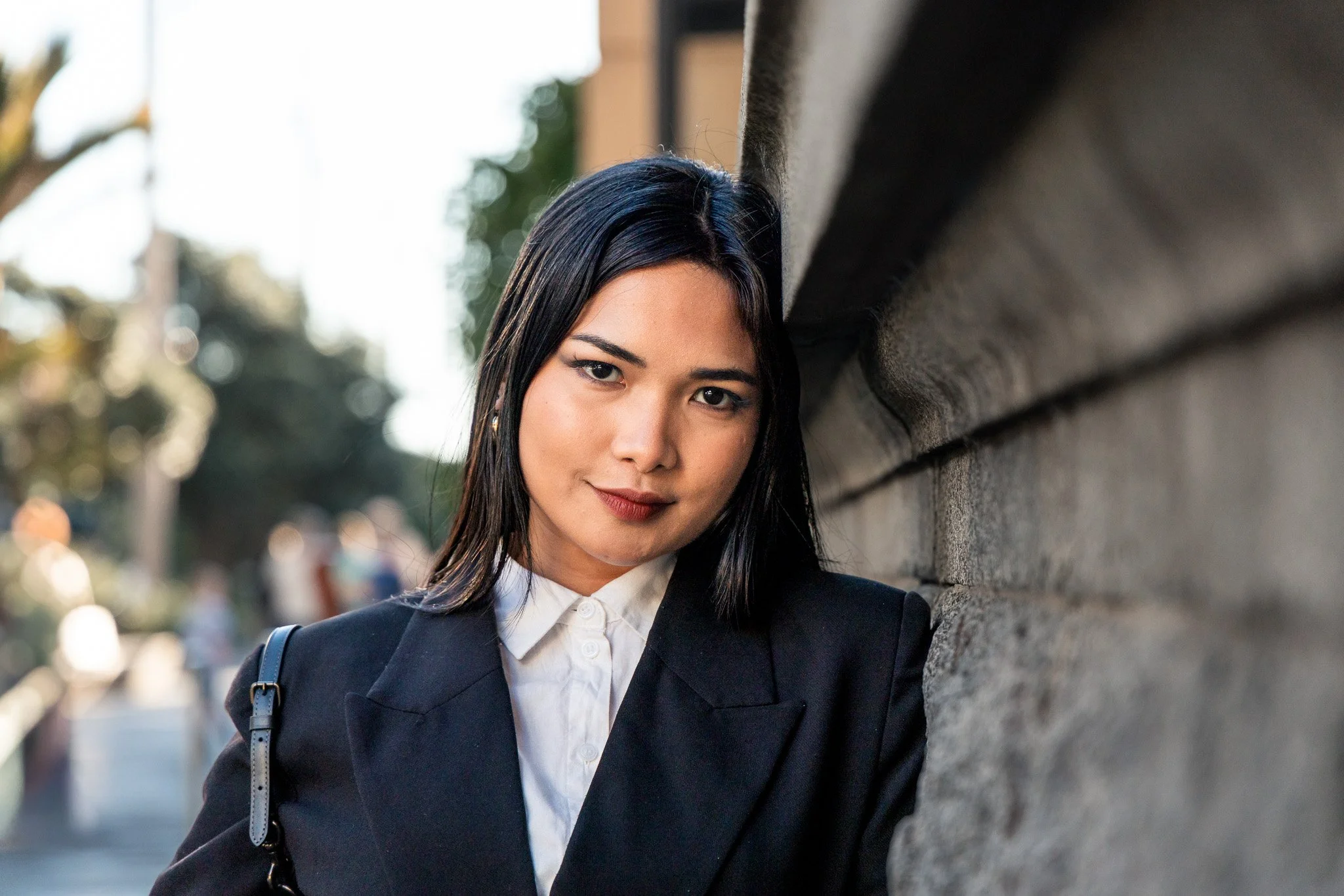 A young woman with long dark hair wearing a black blazer, white shirt, and carrying a black bag, leaning against a stone wall outdoors in a city environment with blurred background of trees and cityscape.