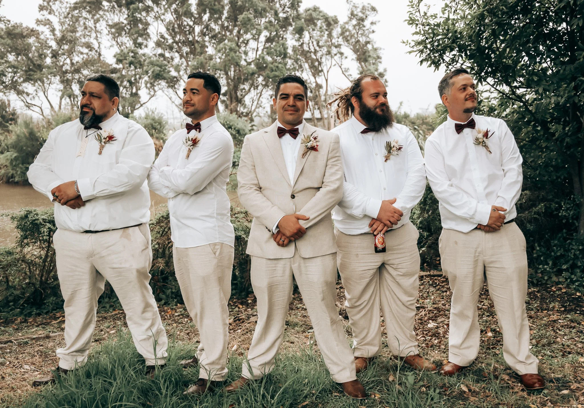 Group of five men dressed in cream suits and white shirts, with three wearing burgundy bow ties and two wearing patterned ties, standing outdoors among trees. They are posed in a line for a photo, with some smiling and others looking serious.