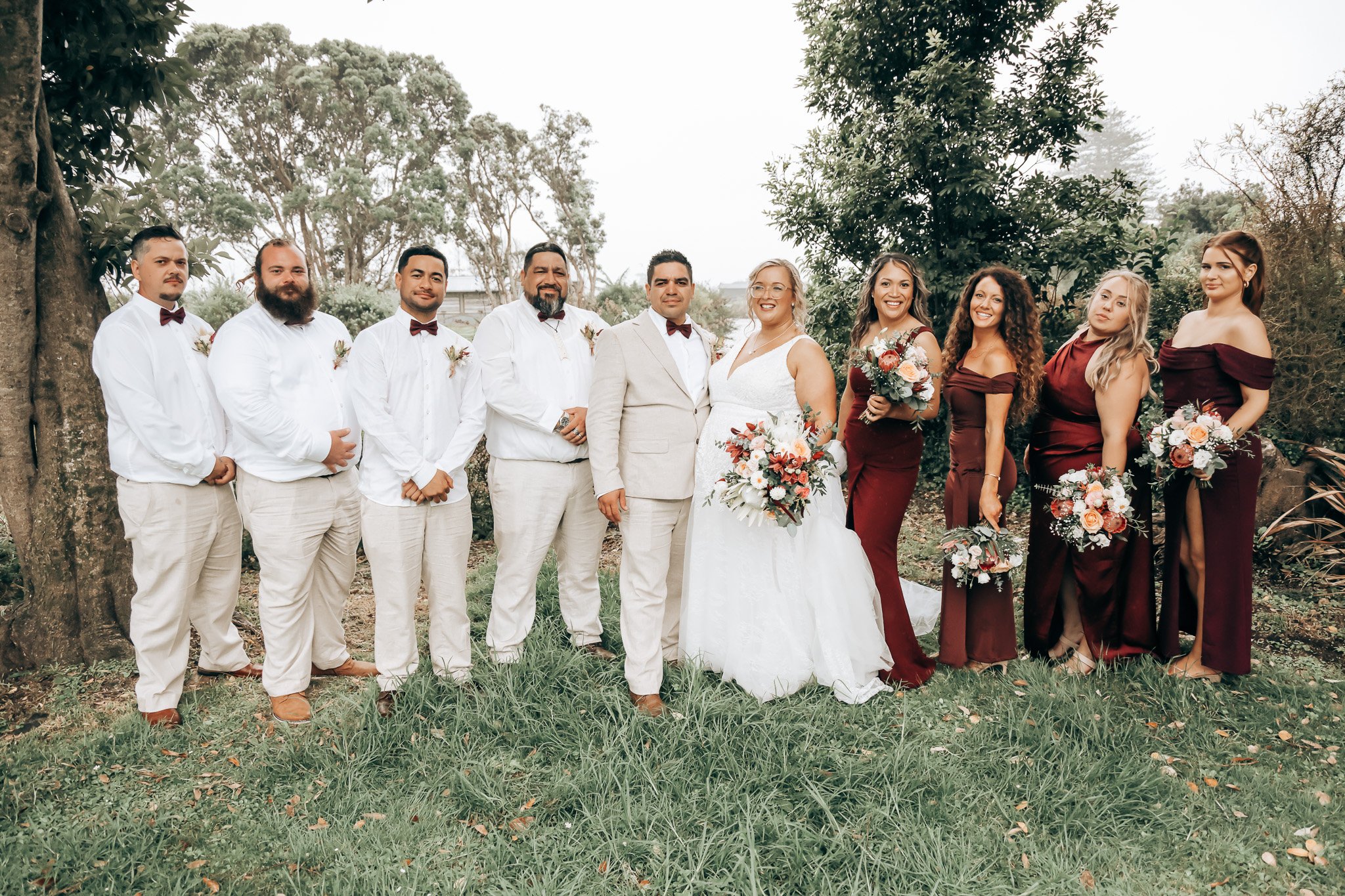 Group of ten people at a wedding, posing outdoors under trees, dressed in formal attire with the bride and groom in the center holding bouquets.