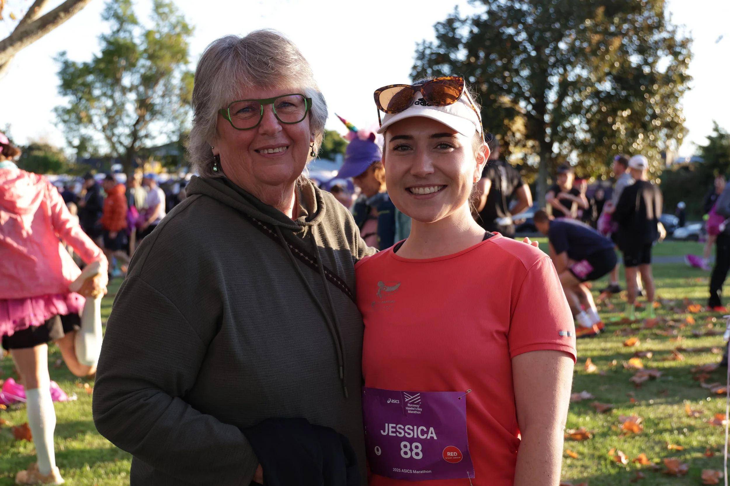 A woman and a young woman are smiling and standing together outdoors during a race event. The young woman is wearing a running bib with the name Jessica, a white visor with sunglasses on top, and a pink shirt. The background shows other participants,