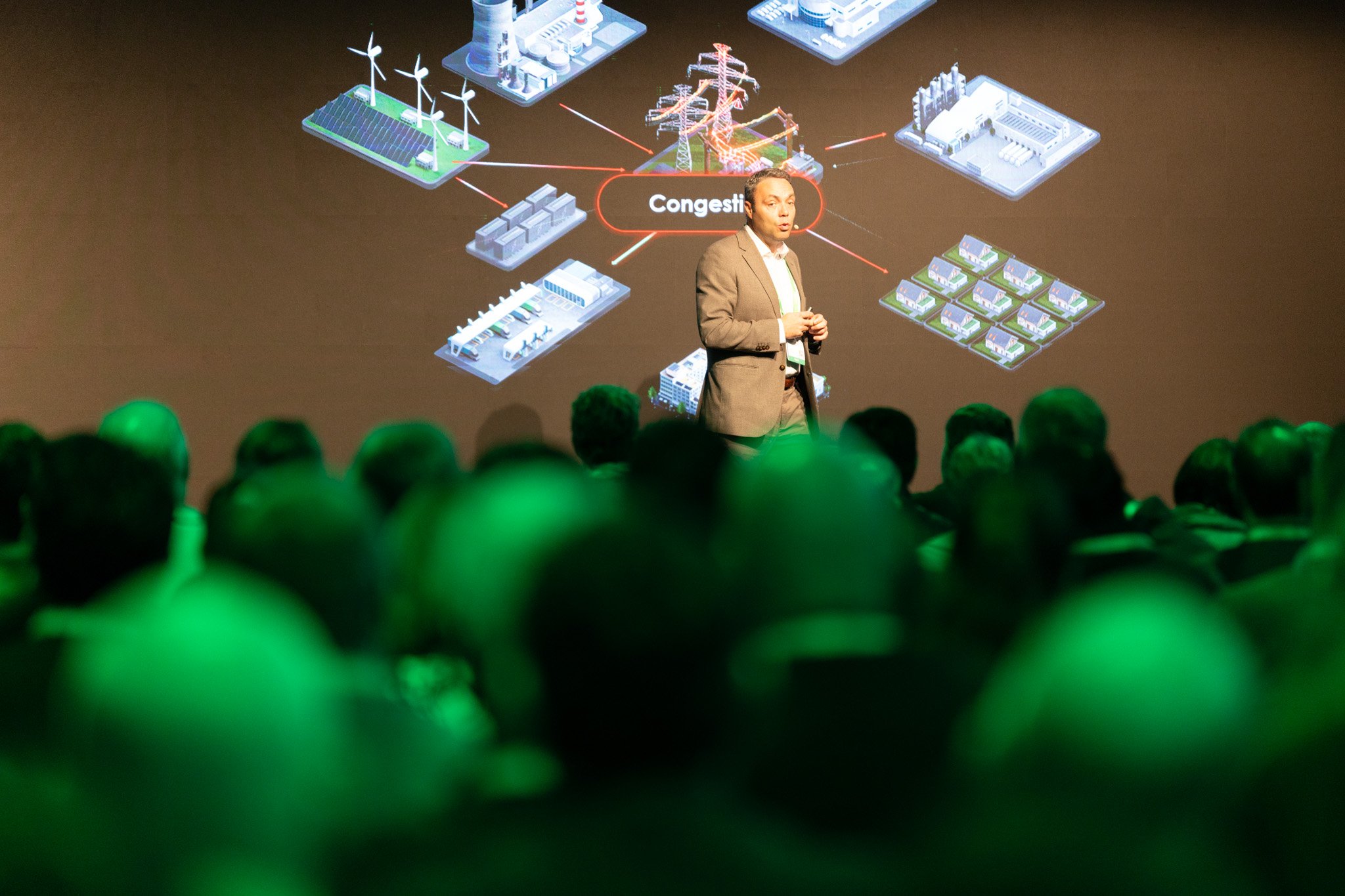 A man giving a presentation on energy grid congestion, with a projected slide showing power plants, wind turbines, solar panels, and electrical infrastructure.