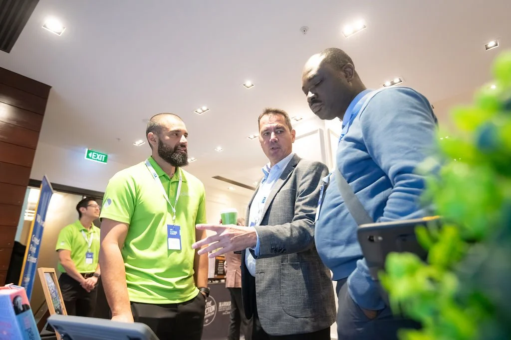 Three men are having a discussion at a conference or event. One man in a green shirt is speaking with two other men, one in a gray suit and the other in a blue hoodie. In the background, another person in a green shirt is standing near a display.