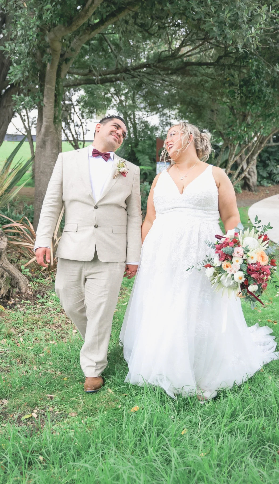 A bride and groom holding hands and smiling at each other outdoors near trees, with green grass and foliage around, during a wedding.