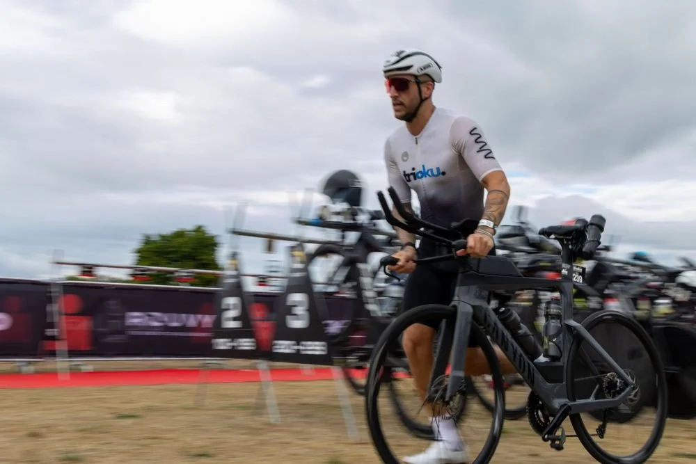 Man wearing a white helmet, sunglasses, and a white triathlon suit riding a black bike during a race.