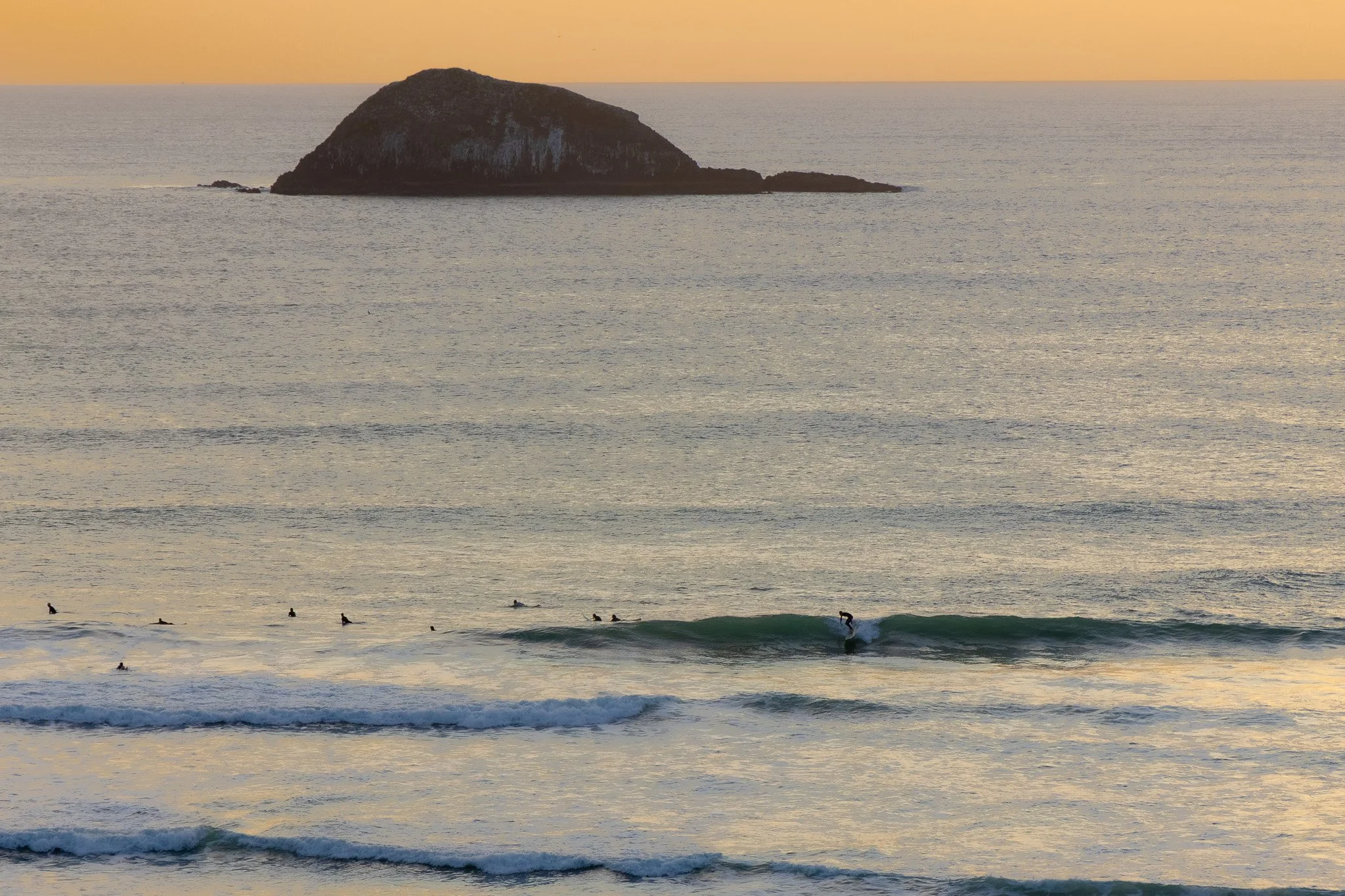 Beach scene with several surfers in the water, a foamy wave breaking near the shore, and a large rock formation in the background during sunset.