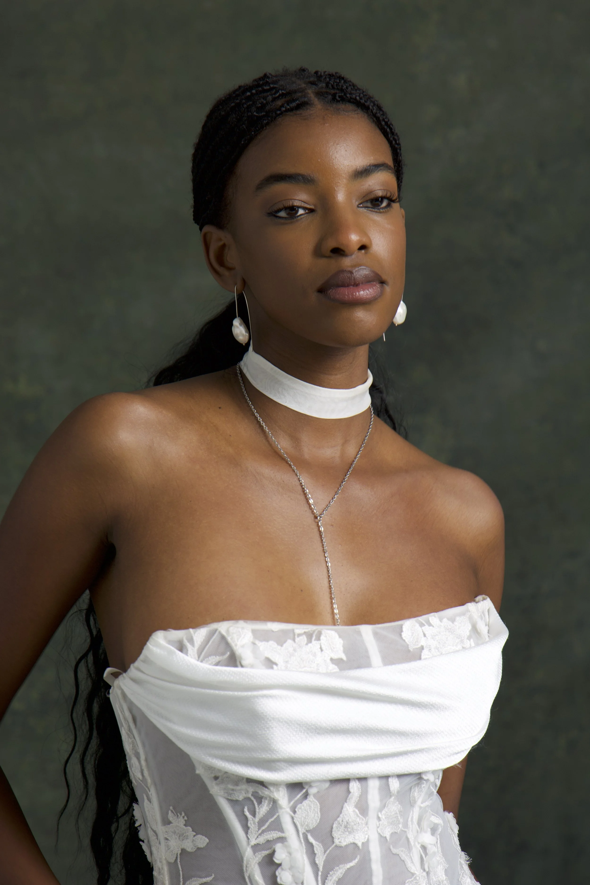 A portrait of a young woman with dark skin, braided hair, and jewelry including earrings, a choker, and a necklace, wearing a white strapless dress with intricate floral lace details, against a plain dark background.