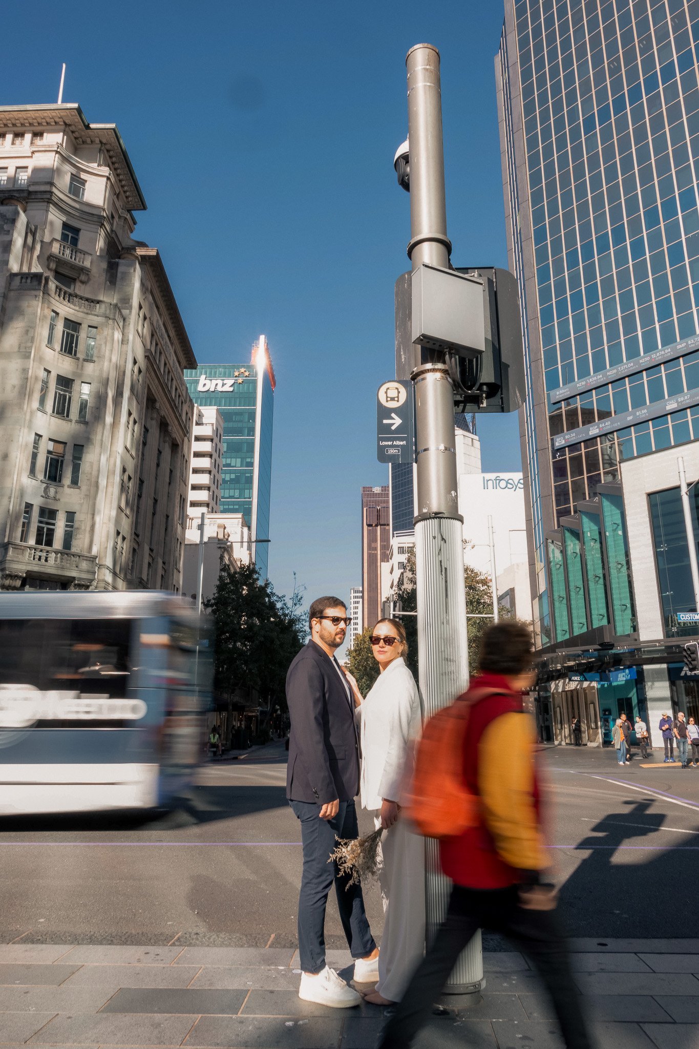 Two people in formal attire, a man and a woman, standing on a city street corner near a pole with a bus stop sign. The cityscape includes tall buildings with glass and stone facades and a blue sky. A bus is blurred in motion on the street.