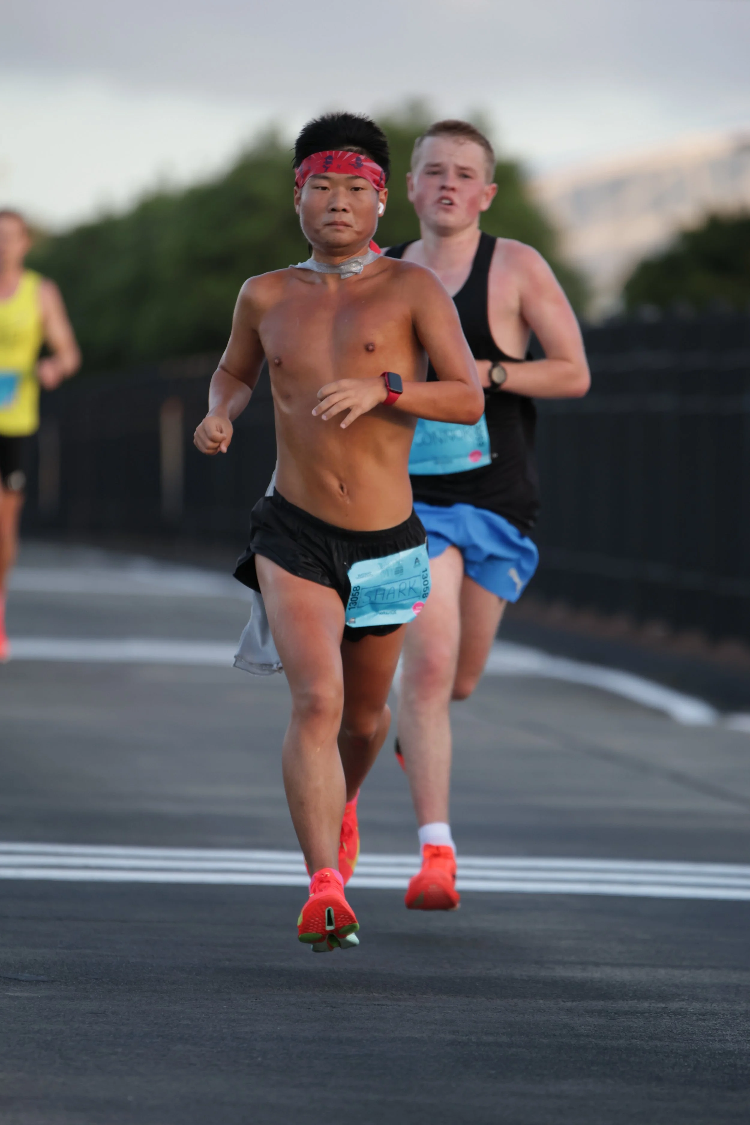 Two male marathon runners running on a road with trees in the background, one shirtless wearing a red headband, black shorts, and red running shoes, the other in a black tank top, blue shorts, and red shoes.