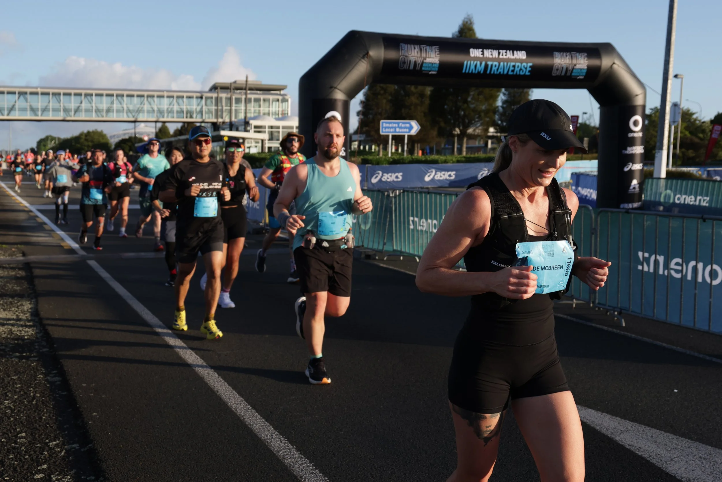 People participating in a marathon race running along a track with a black inflatable arch overhead that reads 'Run the City, One New Zealand, IIKM Traverse.' The front runner is a woman wearing a black tank top, black shorts, a black cap, and a wais