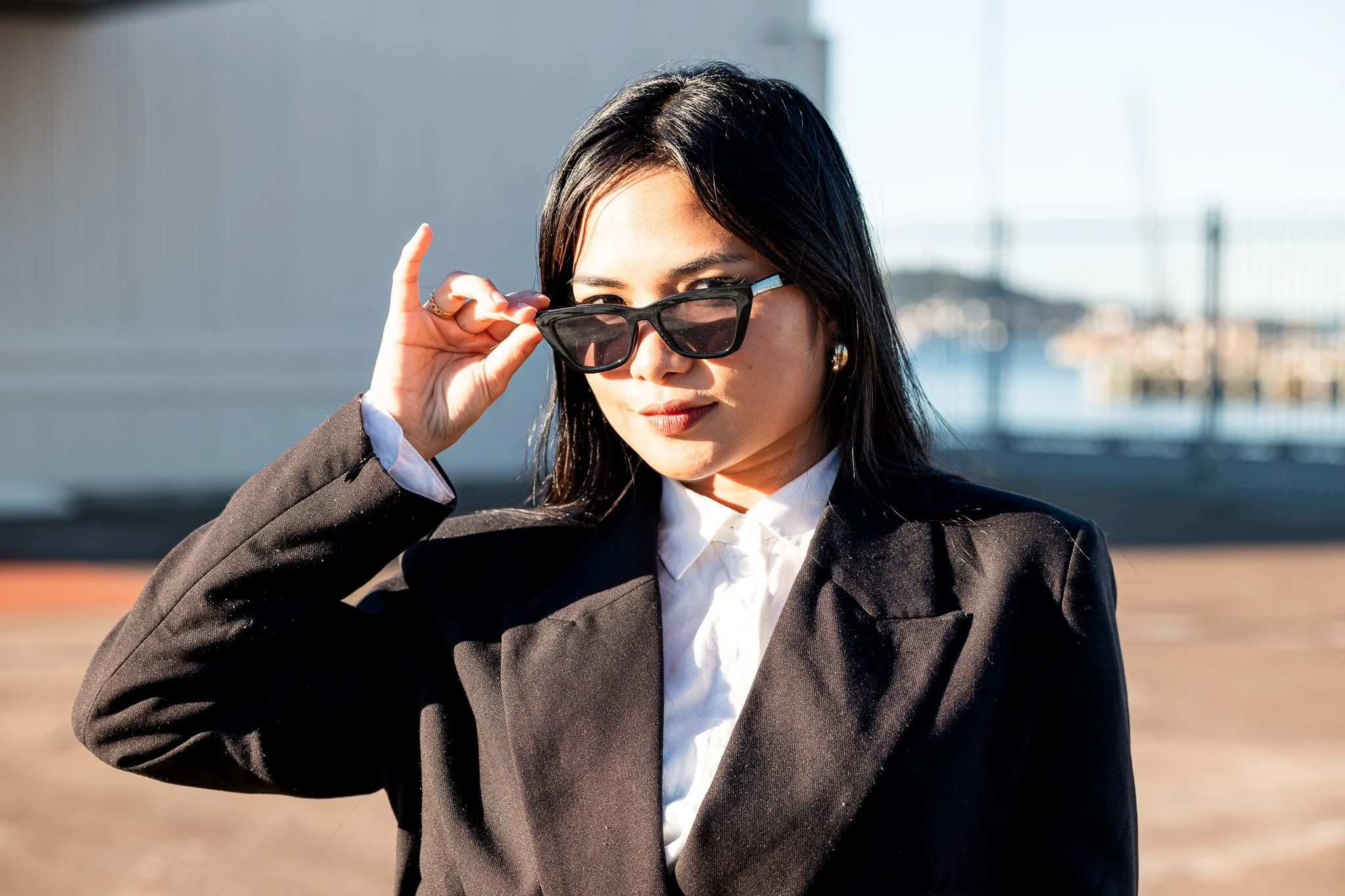 A woman with dark hair wearing black sunglasses and a black blazer, adjusting her sunglasses with her hand, outdoors with blurred background.