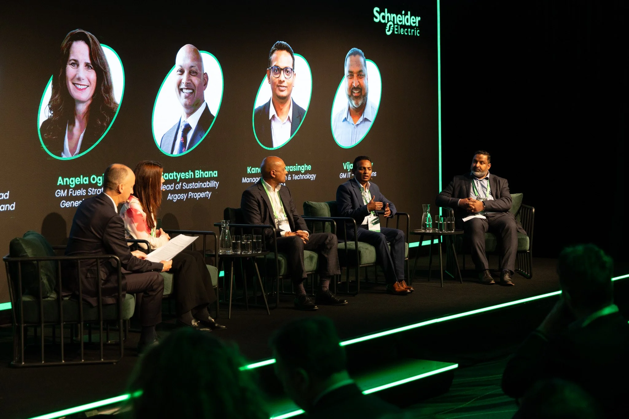 Panel discussion at a conference with six panelists sitting on stage, a large screen displaying portraits and names of the speakers, and a dark background with glowing green accents.