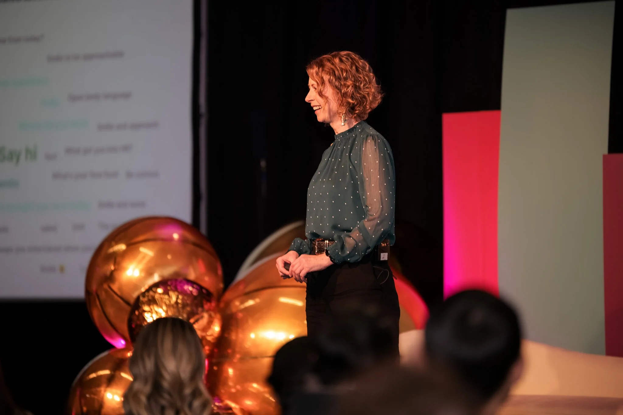 A woman with curly blonde hair smiling while speaking at an event, standing in front of large gold balloons and a colorful background.