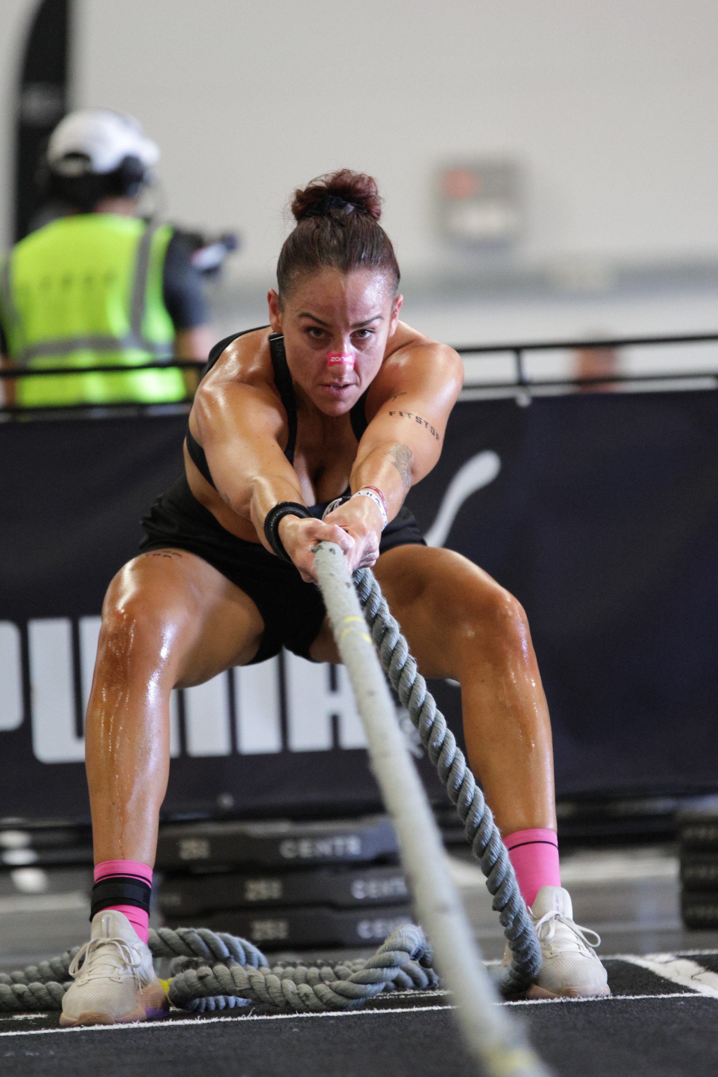 Female athlete in black workout attire, pink socks, and white shoes, pulling a thick battle rope during a fitness competition or workout session.