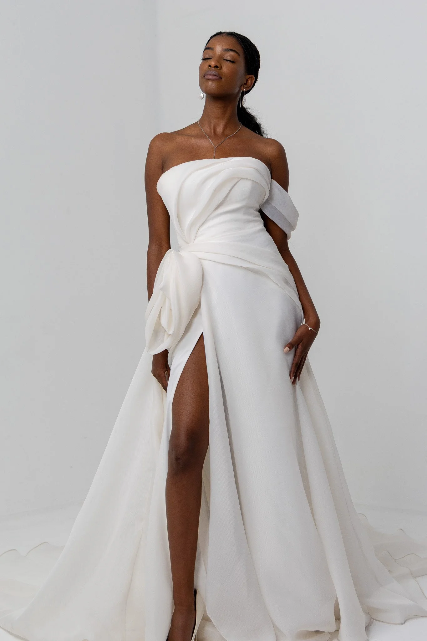 A woman in a white off-shoulder gown with a thigh-high slit, standing against a plain light gray background, with her eyes closed and a serene expression.