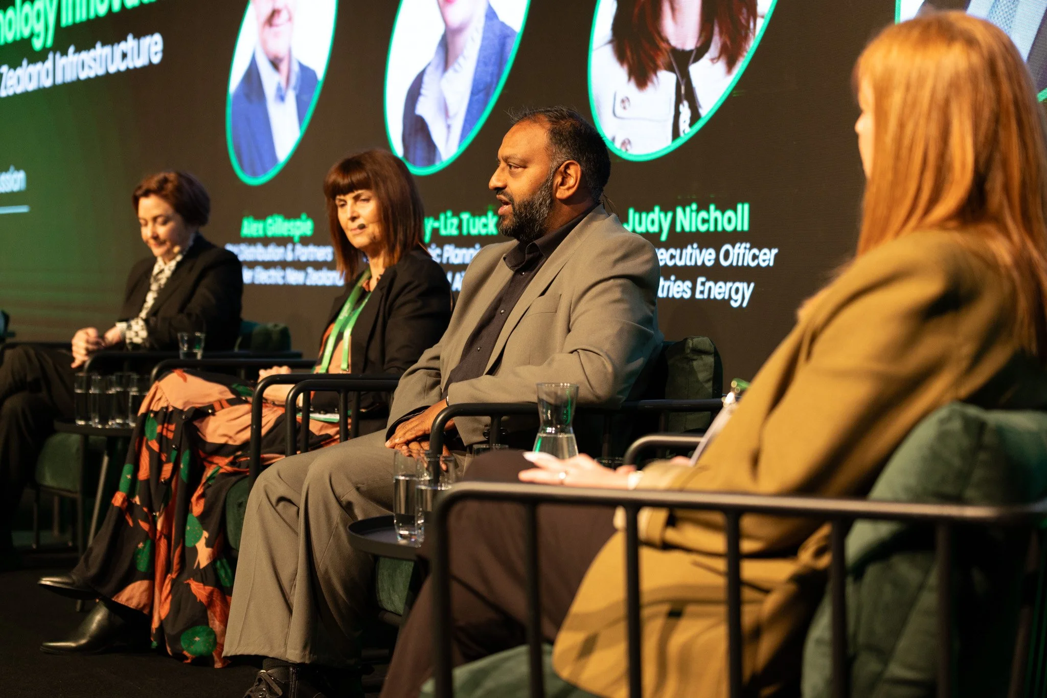 Four panelists sitting on stage at a conference, engaging in discussion. Background displays highlighted names and titles, including Judy Nicholl, with images of individuals. The panelists include women and men dressed professionally, with water glas
