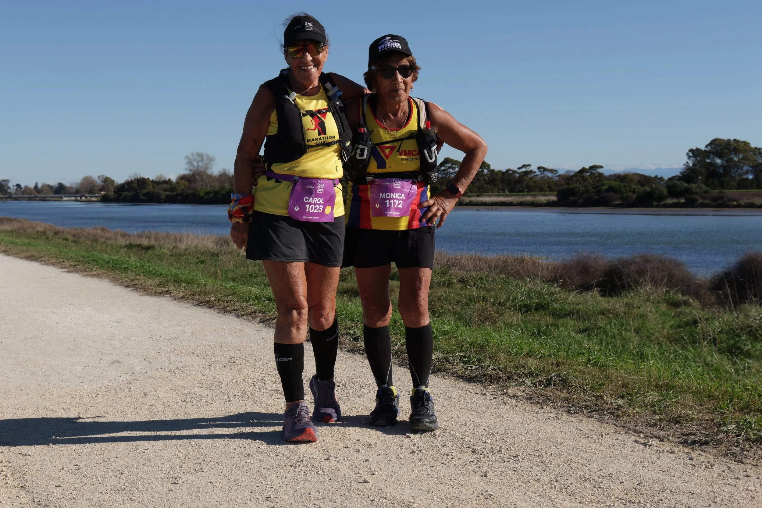 Two women in athletic gear standing on a dirt path near a body of water, with trees and clear blue sky in the background.