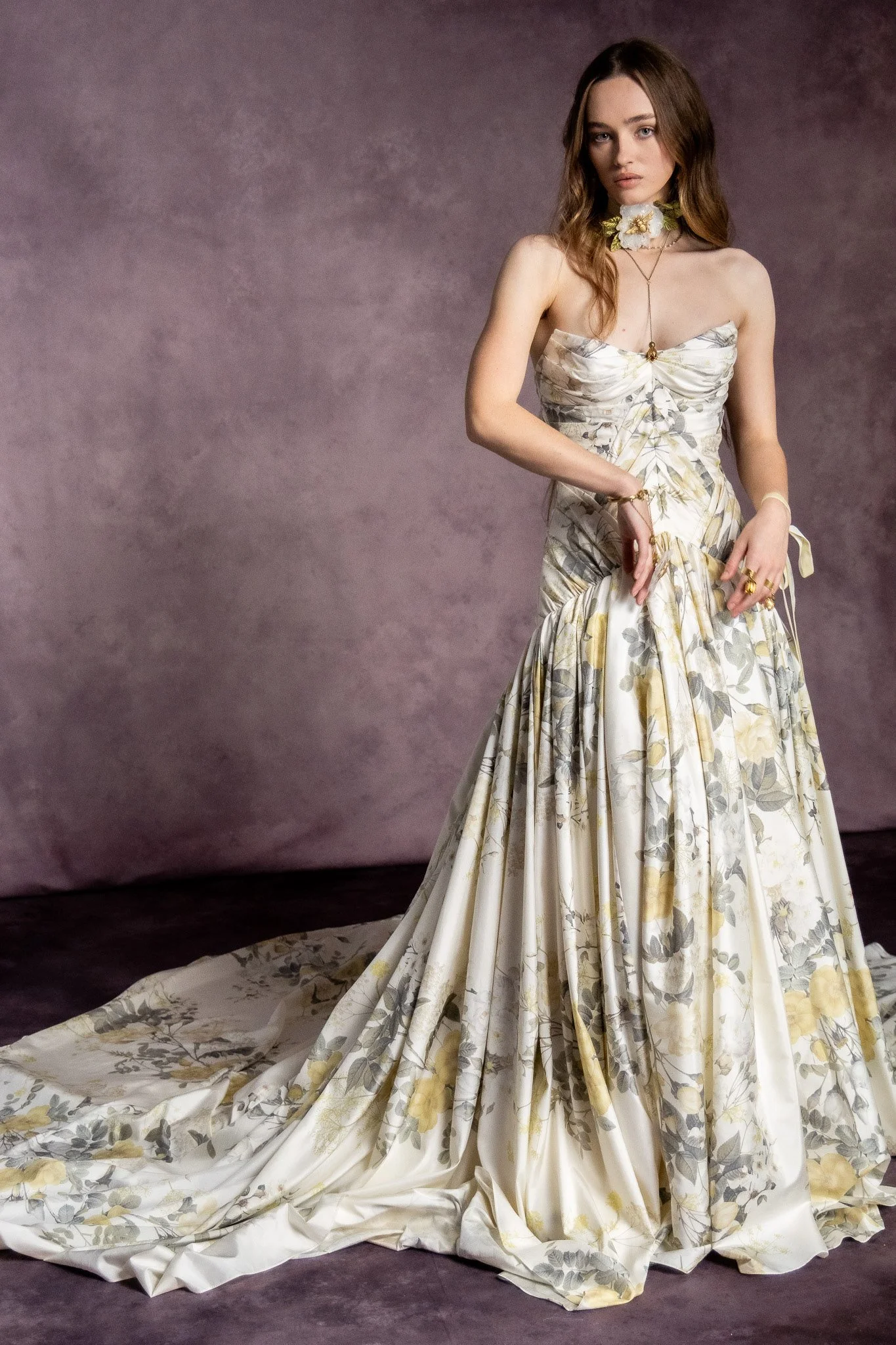A woman with long brown hair wearing a strapless floral gown with a long train, standing in front of a textured purple background.