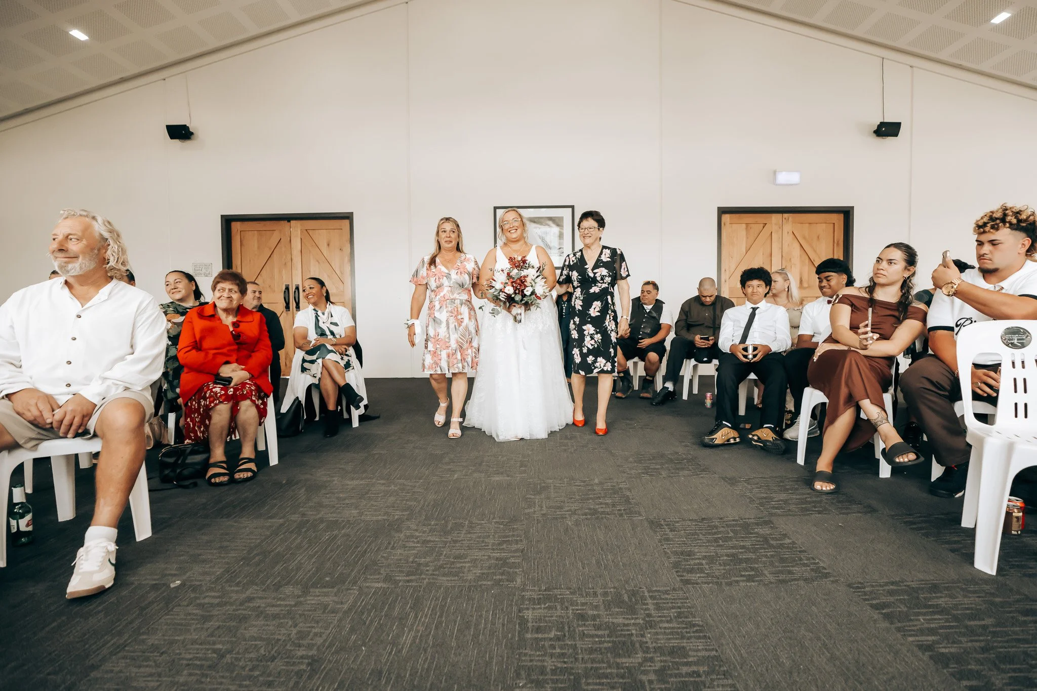 A bride in a white wedding dress and a bouquet of flowers walking down the aisle, accompanied by two women, in a room filled with seated guests.