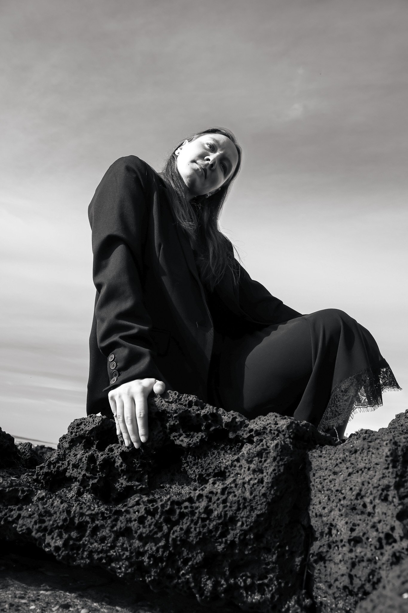 Black and white photo of a woman sitting on a rocky surface, wearing a black jacket and a long skirt, with the sky in the background.