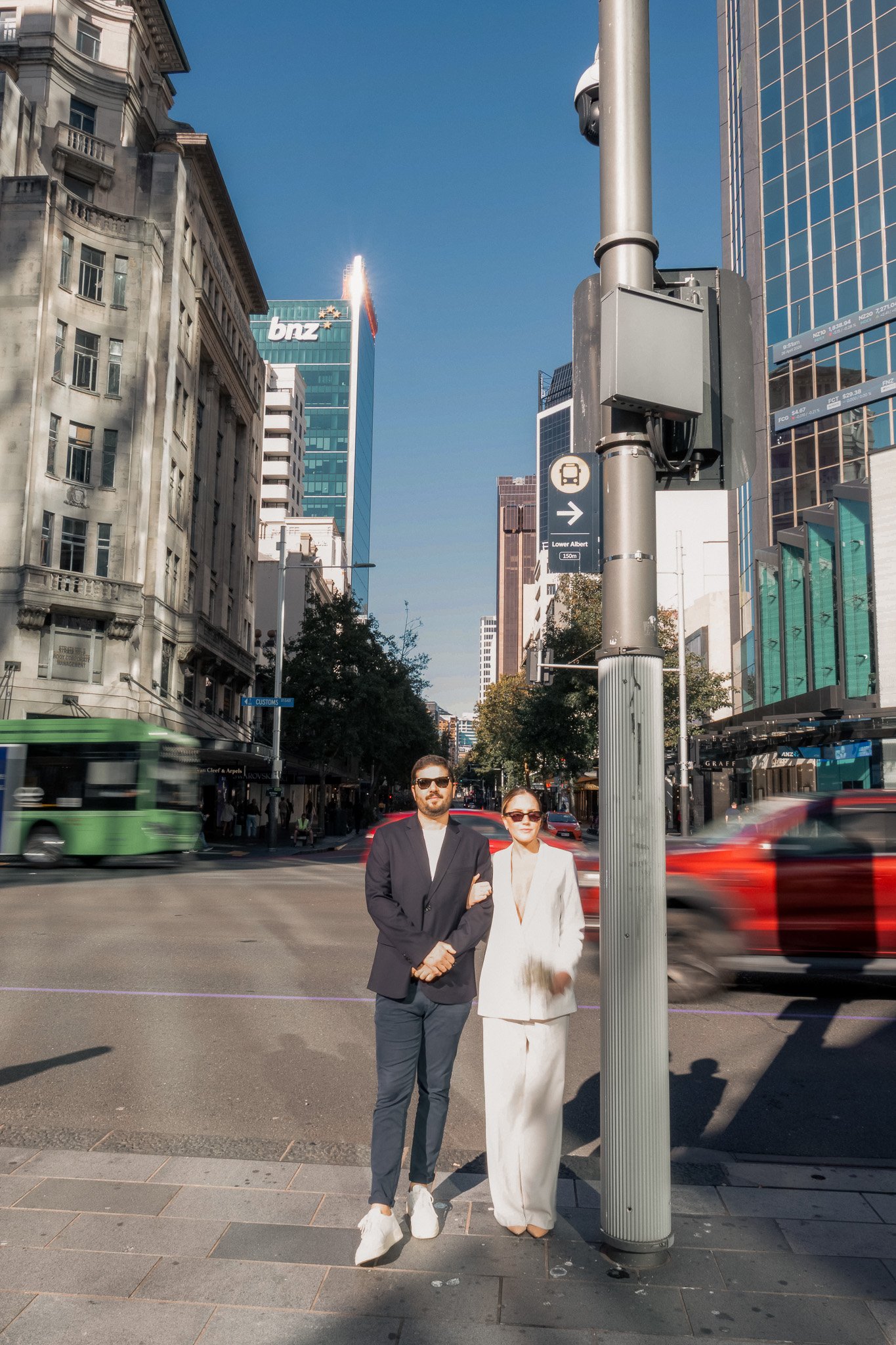 Two people, a man and a woman, standing on a city street corner with tall buildings and moving vehicles in the background, in bright daylight.