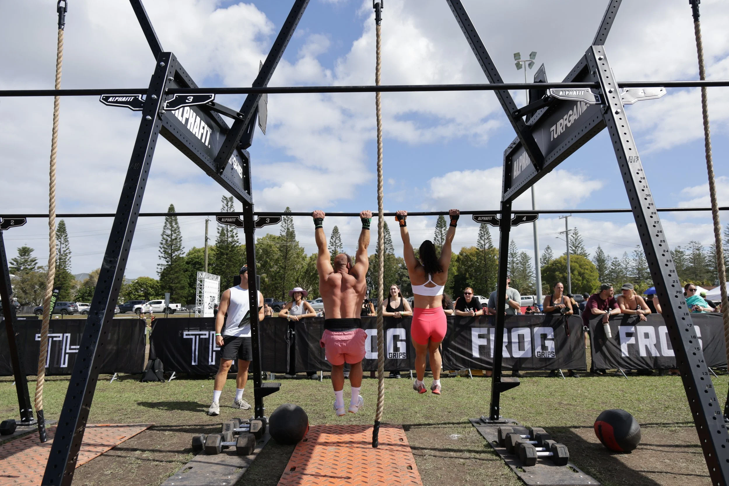 Two men are hanging from pull-up bars at an outdoor fitness event. Spectators watch behind a black barrier, and there are trees, cars, and a cloudy sky in the background.