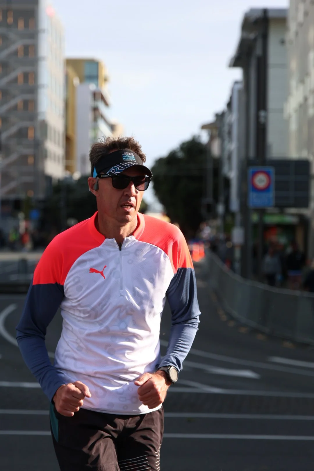 A man running in an urban area, wearing sunglasses, a headband, a Puma sports shirt, and a watch, with tall buildings in the background.