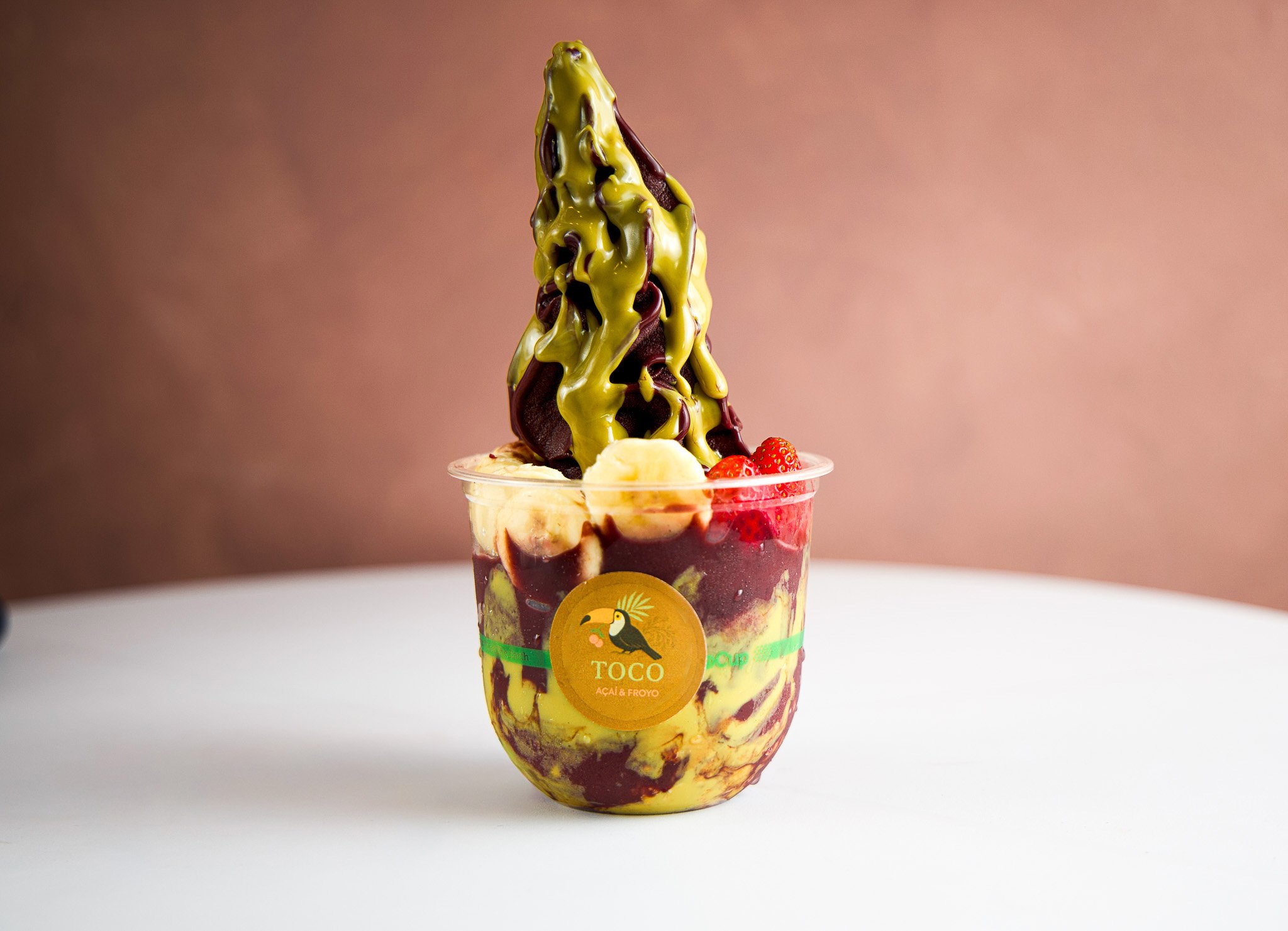 A cup of frozen dessert called açaí and froyo with a tall, twisted topping of dark syrup and yellow sauce, garnished with strawberries, banana slices, and a straw.