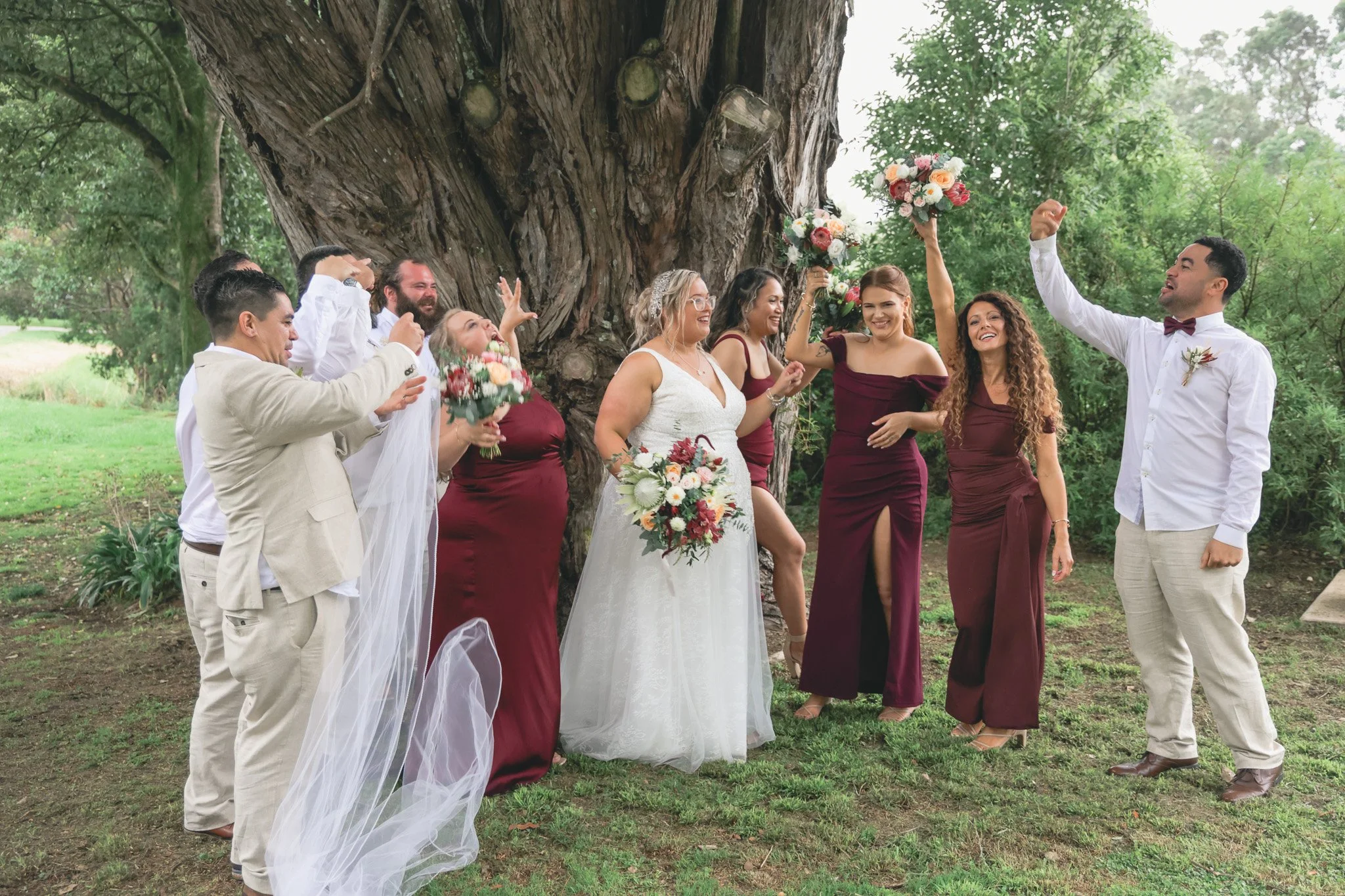 A wedding celebration outdoors with a bride, groom, and bridesmaids surrounded by a large tree and greenery. The bride is holding a bouquet and wearing a white wedding dress, and the groom is wearing a white shirt with a bow tie. Some bridesmaids are