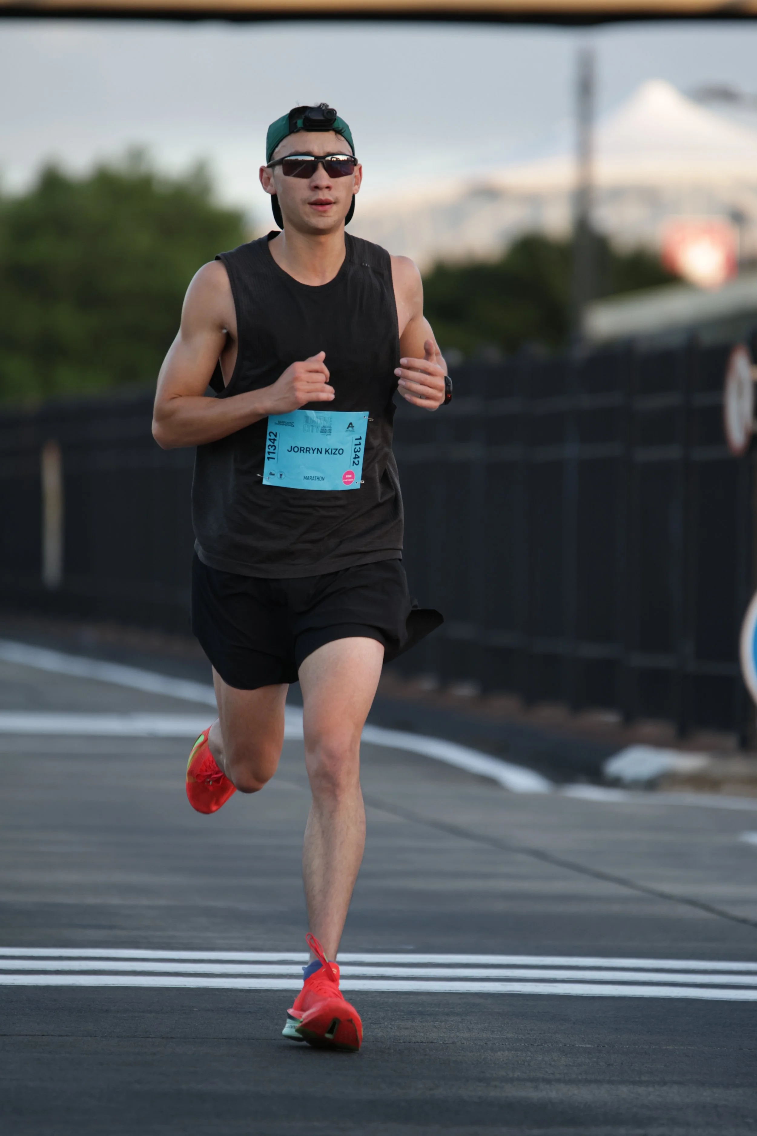 Male runner in black athletic wear, sunglasses, and a green cap running on a road during the daytime.