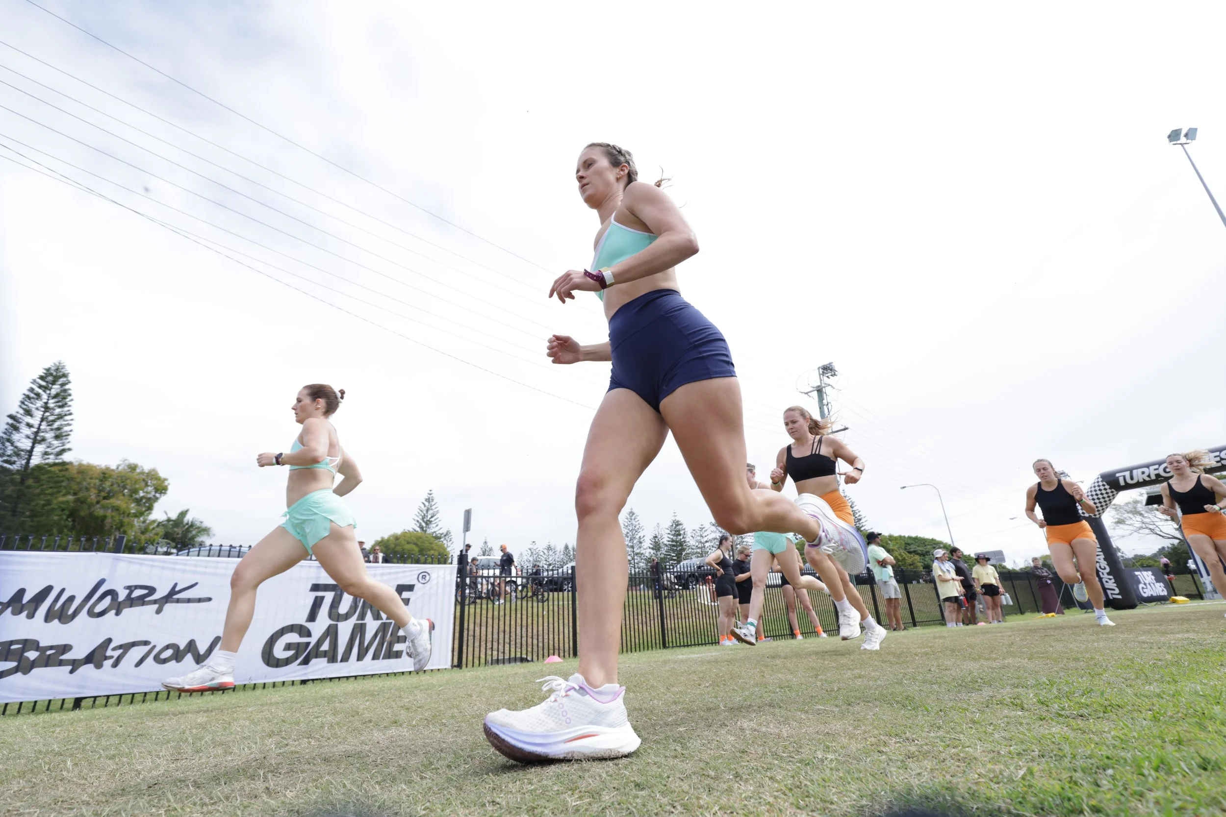 Women running on a track during a race at an outdoor athletic event with a crowd and banners in the background.