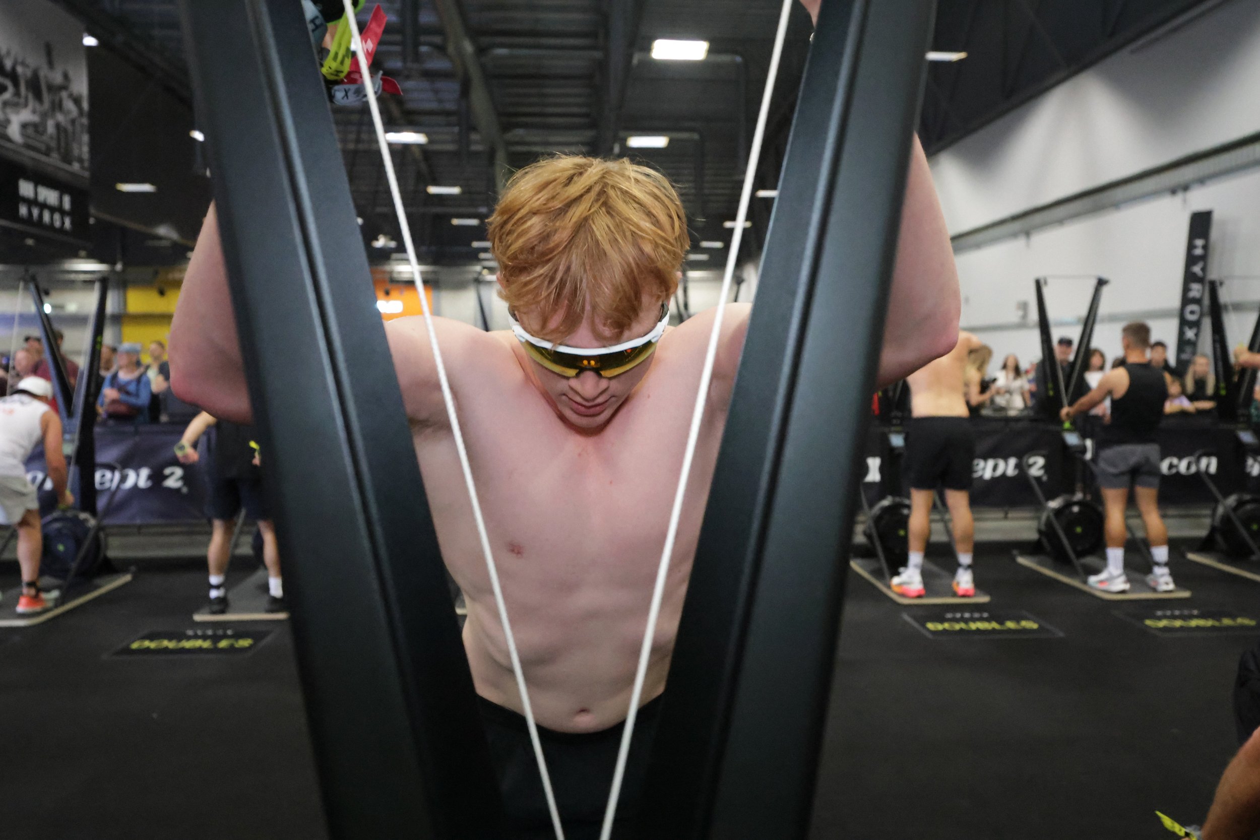A shirtless man with red hair, wearing sunglasses, is doing an overhead stretch in a gym. He is surrounded by people working out in the background.