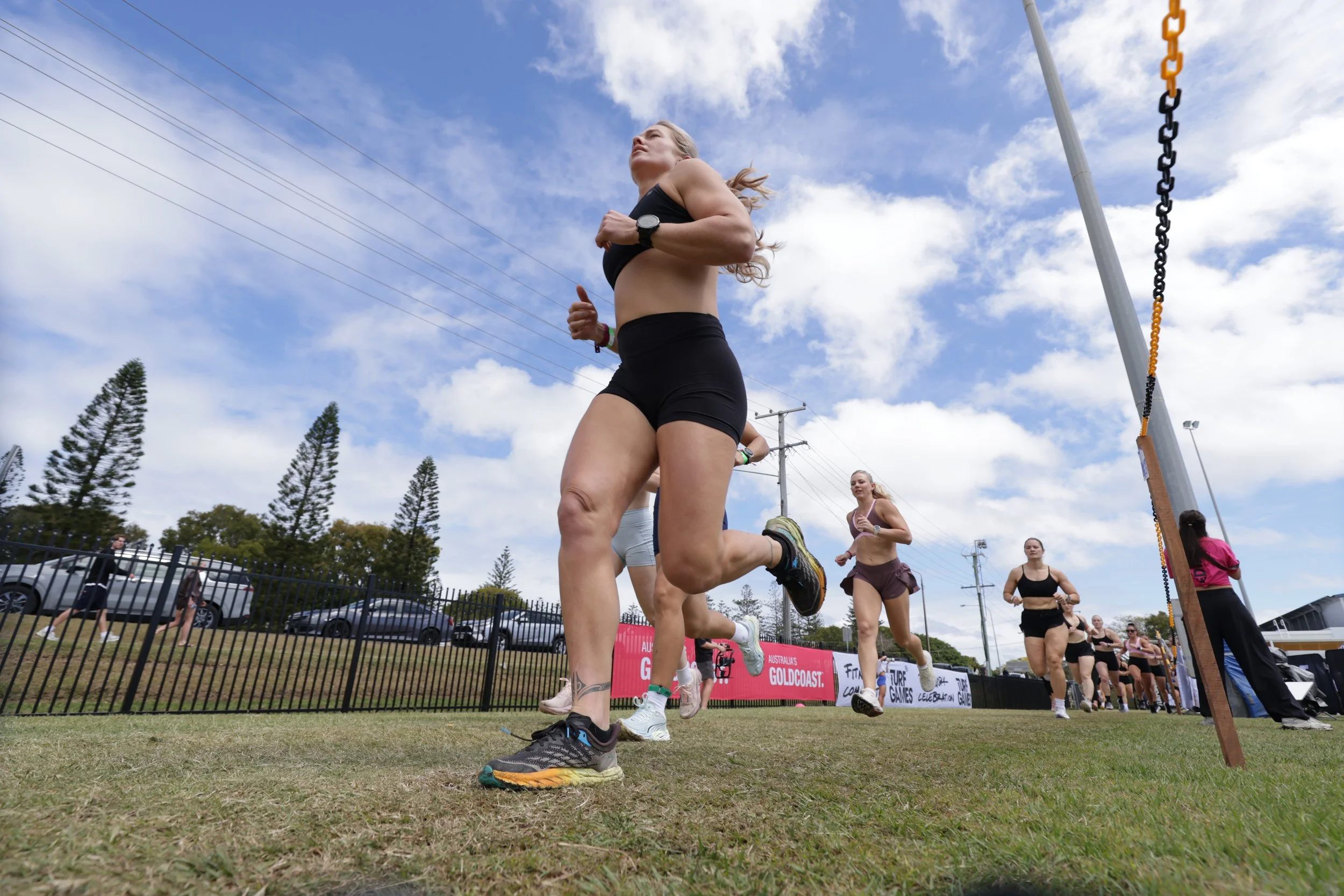 Women participating in a running race outdoors on a cloudy day. Several women are running in a line, with tall trees and parked cars visible in the background. The runners are dressed in athletic clothing and running shoes.