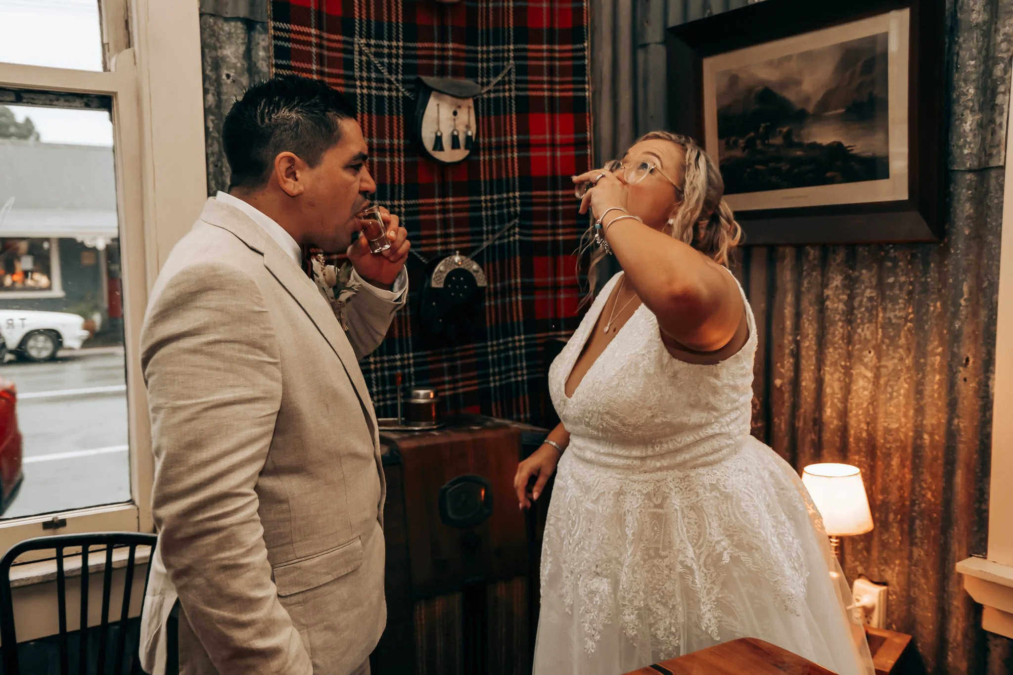A man in a light-colored suit and a woman in a white lace wedding dress are inside a warmly lit room. The man is drinking a shot glass, and the woman is holding her hand to her face, wearing glasses. The background features a patterned tartan wall ha