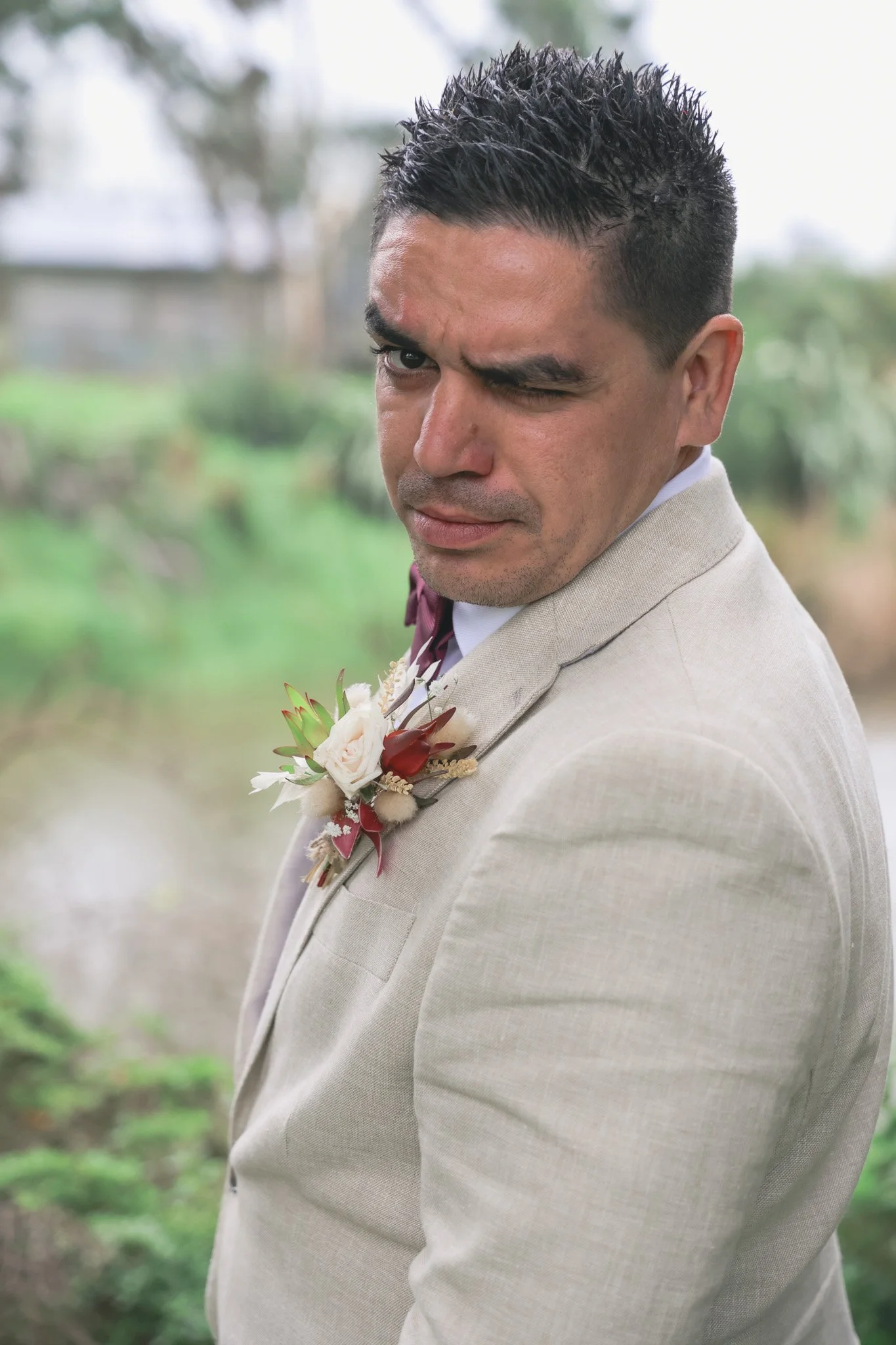 A man in a light-colored suit with a pink and white boutonniere, winking at the camera outdoors with blurred greenery in the background.