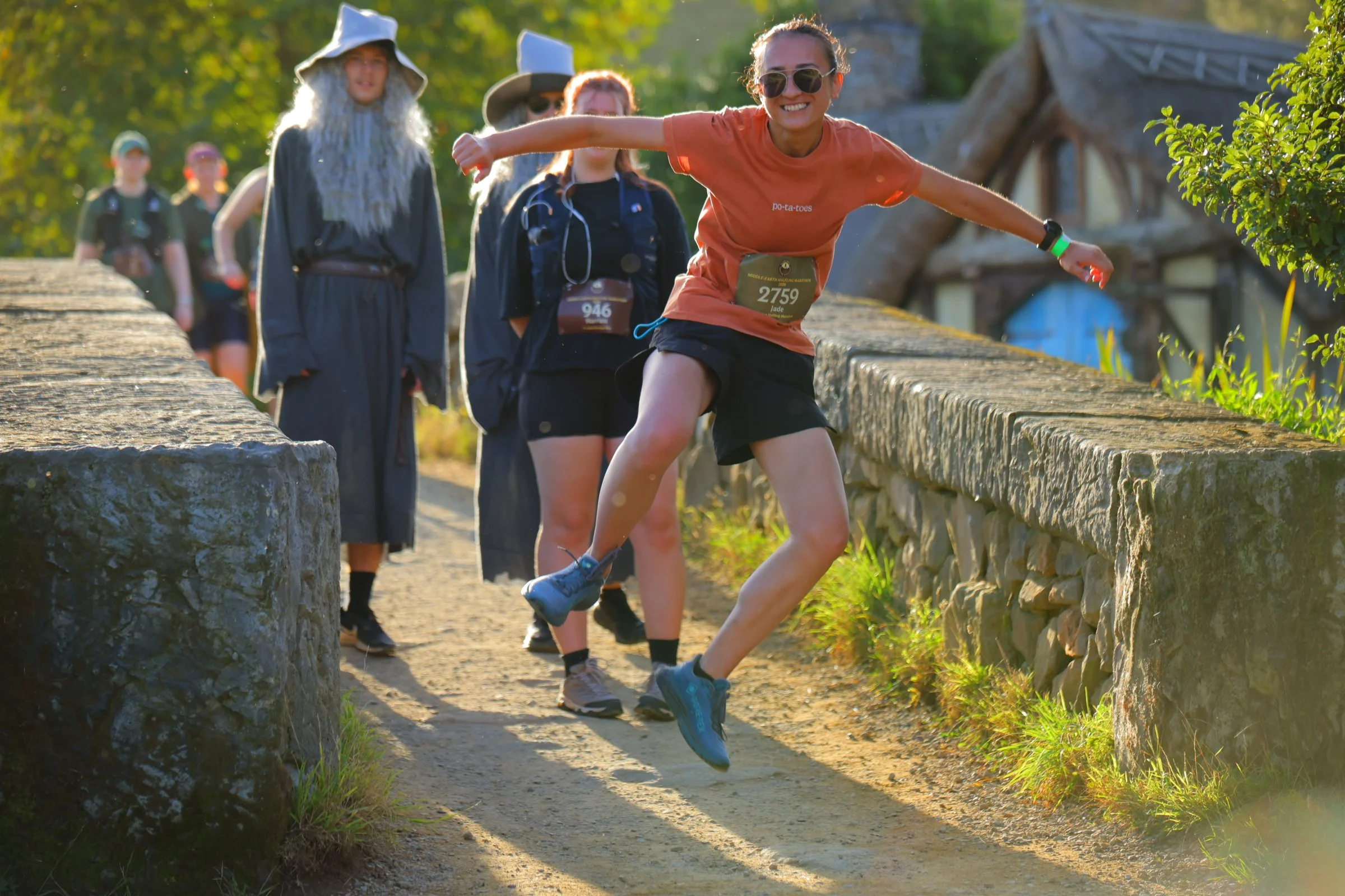 A young woman running and jumping on a dirt trail with a stone wall on one side, during a race or outdoor event, with other participants and greenery in the background.