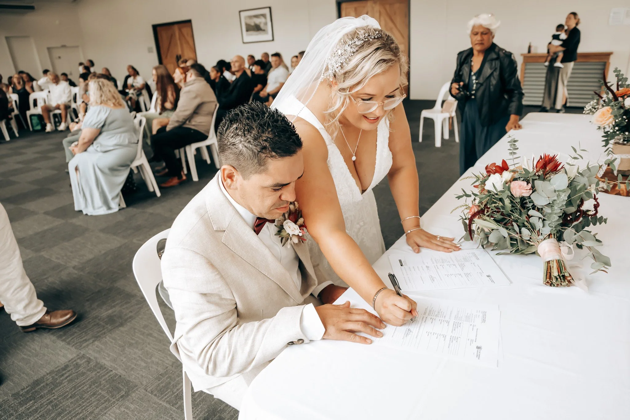 A bride and groom signing documents at a wedding reception. The bride is wearing a white wedding dress and veil, and the groom is dressed in a light-colored suit. Guests are seated in the background, and floral decorations are on the table.