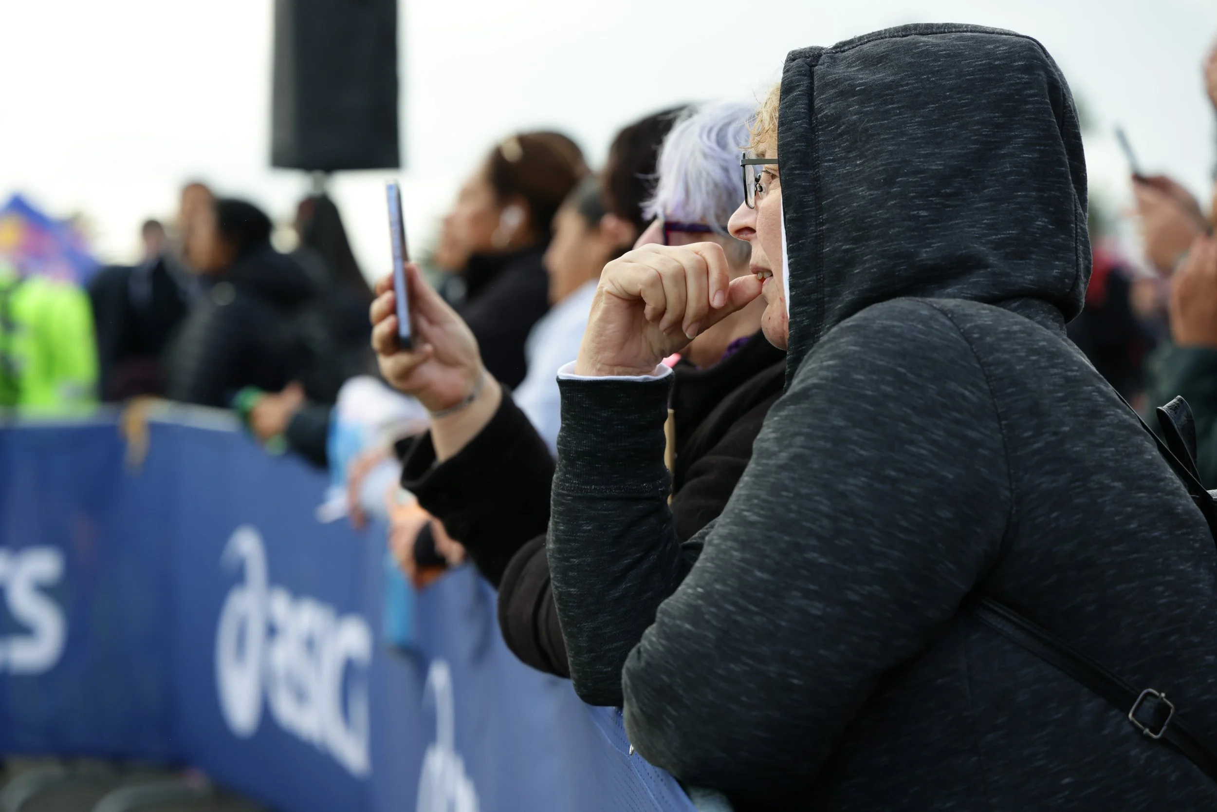 A group of people seated at an outdoor event, with one woman in a black hoodie and glasses in the foreground, holding her hand to her mouth, while others behind her are watching, some holding their phones.