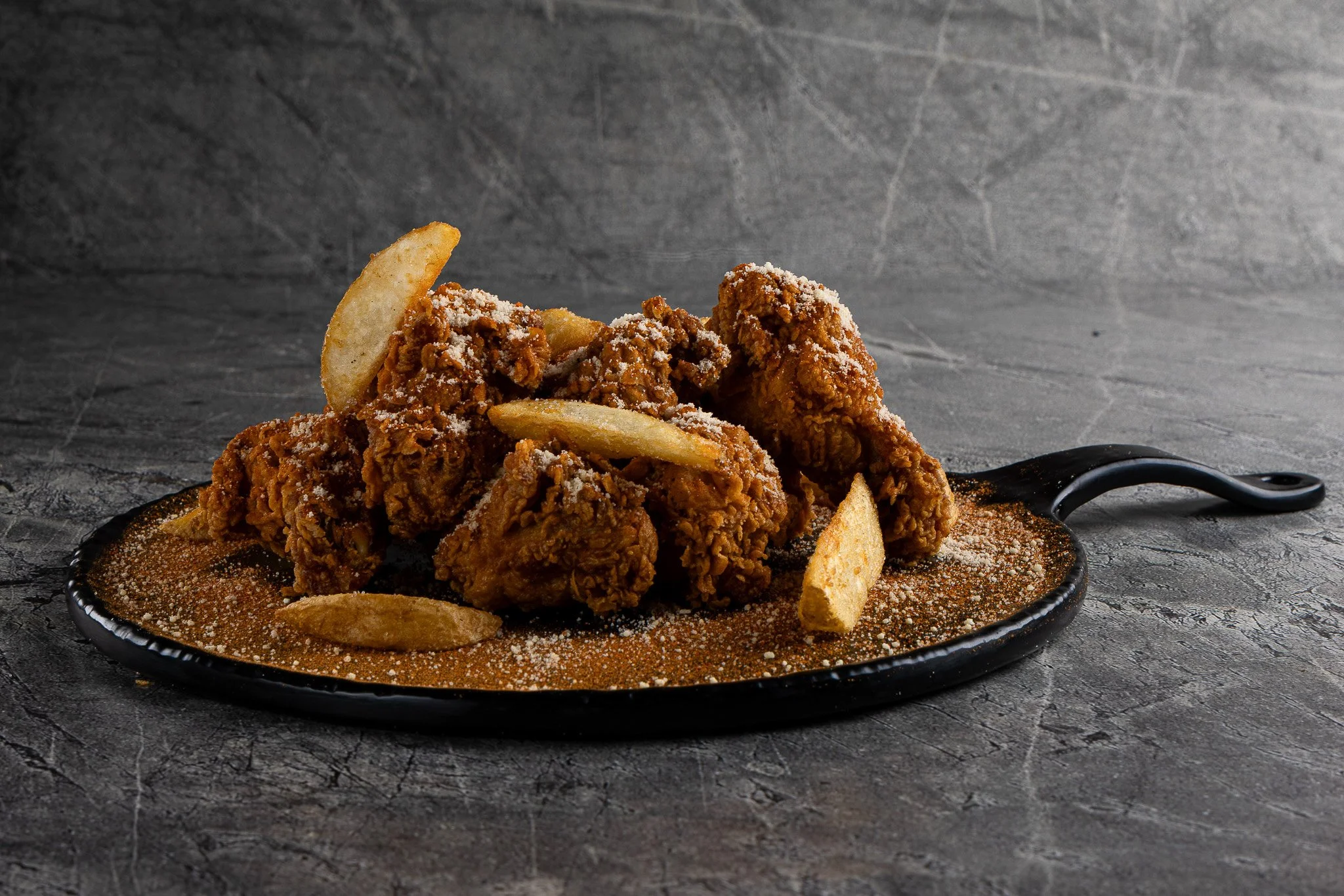 Fried chicken pieces with potato chips on a black plate with red seasoning, on a dark textured surface.