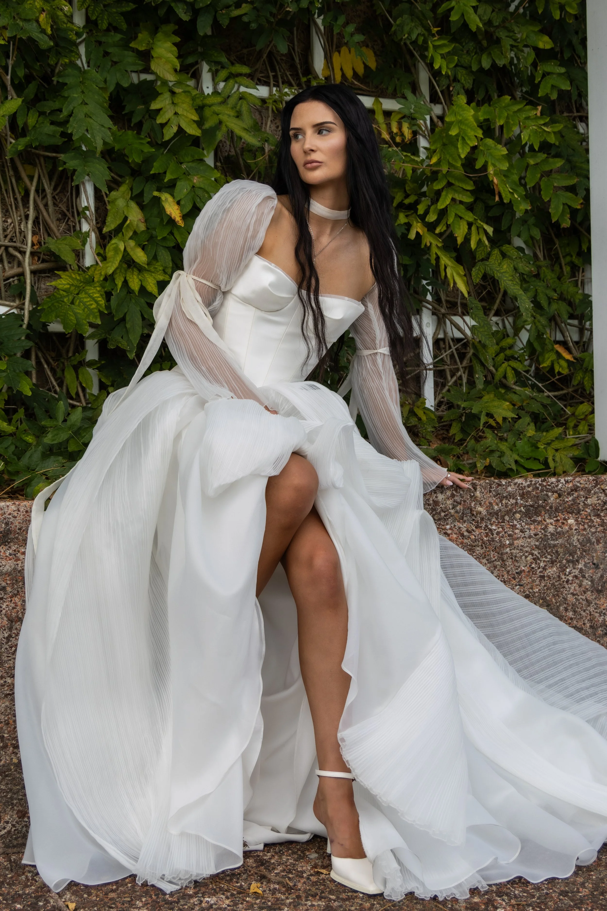 A woman with long black hair wearing a white flowing dress with sheer puffed sleeves, sitting outdoors against a background of green leaves and vine-covered wall, with one leg exposed and her hand resting on the ground.
