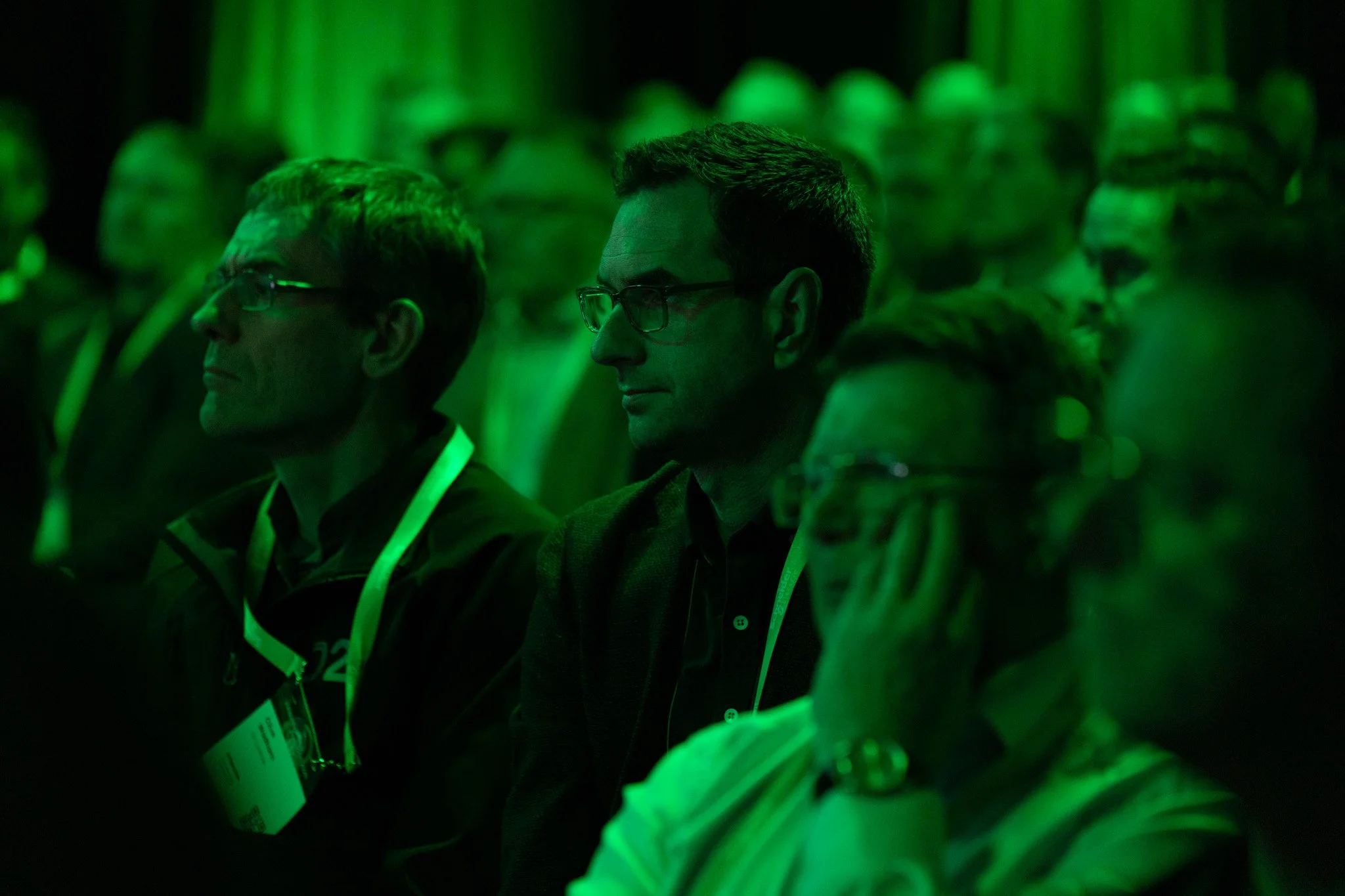 People attending a conference or event, seated and listening, illuminated by green lighting.