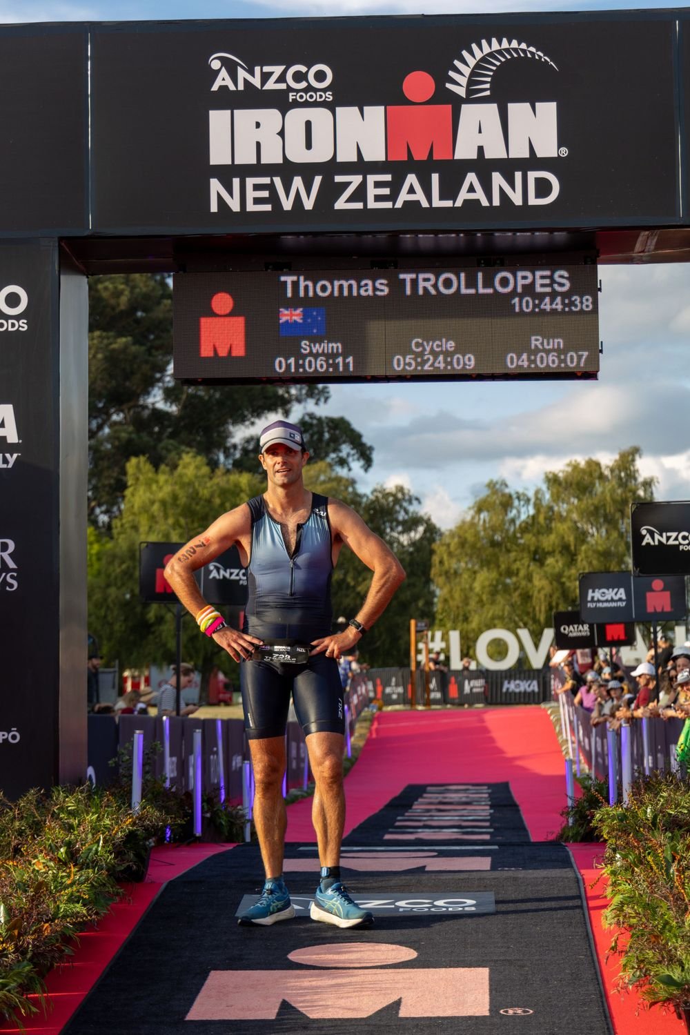 Triathlon athlete standing on the finish line of an Ironman race in New Zealand, with a digital timer and race details above. The athlete is wearing a blue and black triathlon suit, a cap, and accessories, with spectators in the background.