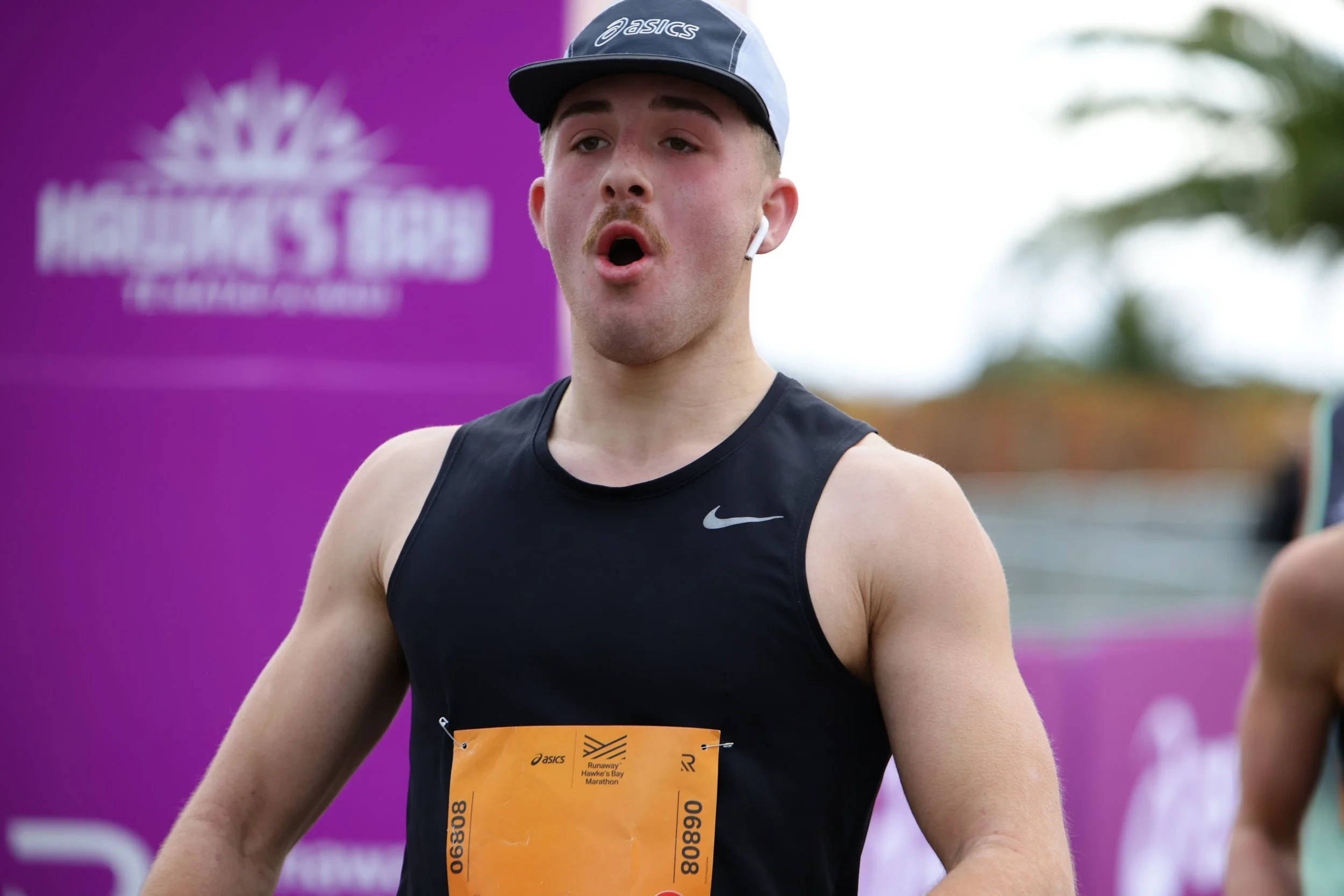 A male athlete wearing a black Nike tank top and a gray cap is participating in a marathon, with a purple event banner in the background. He has a race bib attached to his stomach and appears to be talking or breathing heavily during the race.