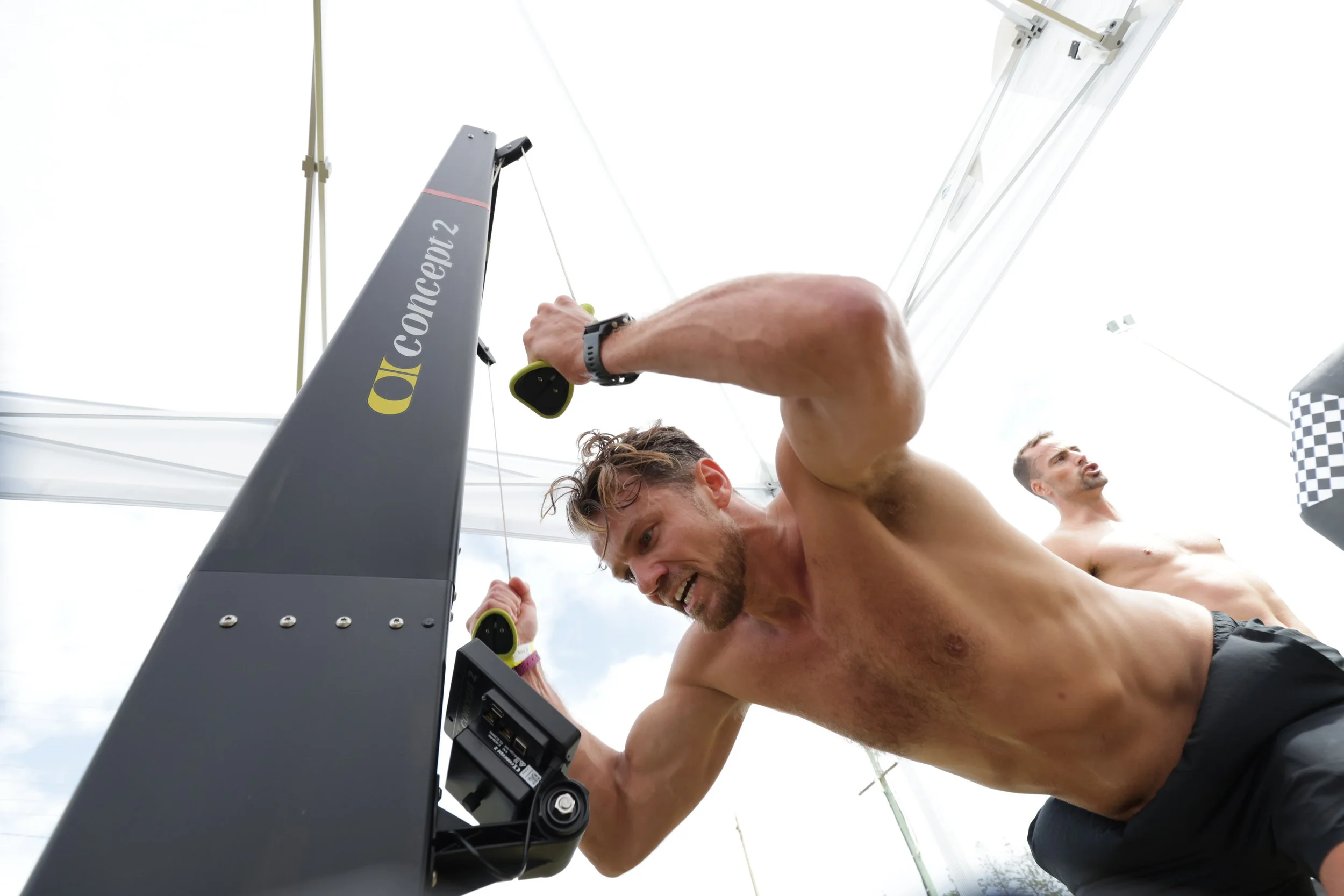 Two shirtless men celebrating after crossing the finish line in a race, with one holding a smartphone and a large sign that reads 'concent 2', under a tent.