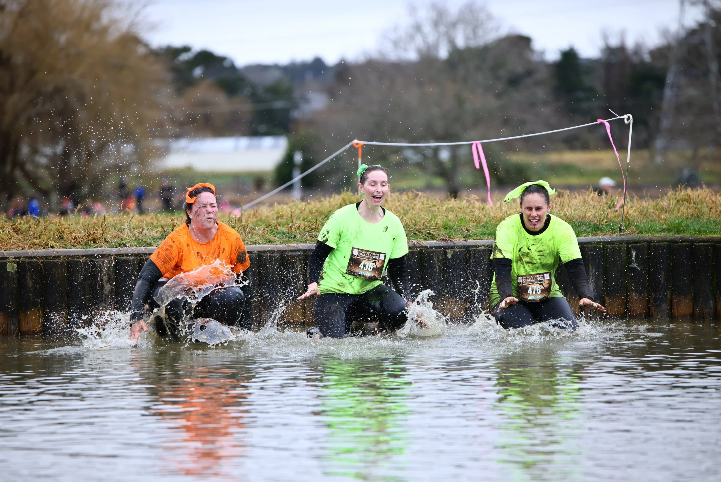 Three women participating in an outdoor obstacle race are wading through a water pit, splashing water as they cross it, wearing brightly colored shirts and race bibs.