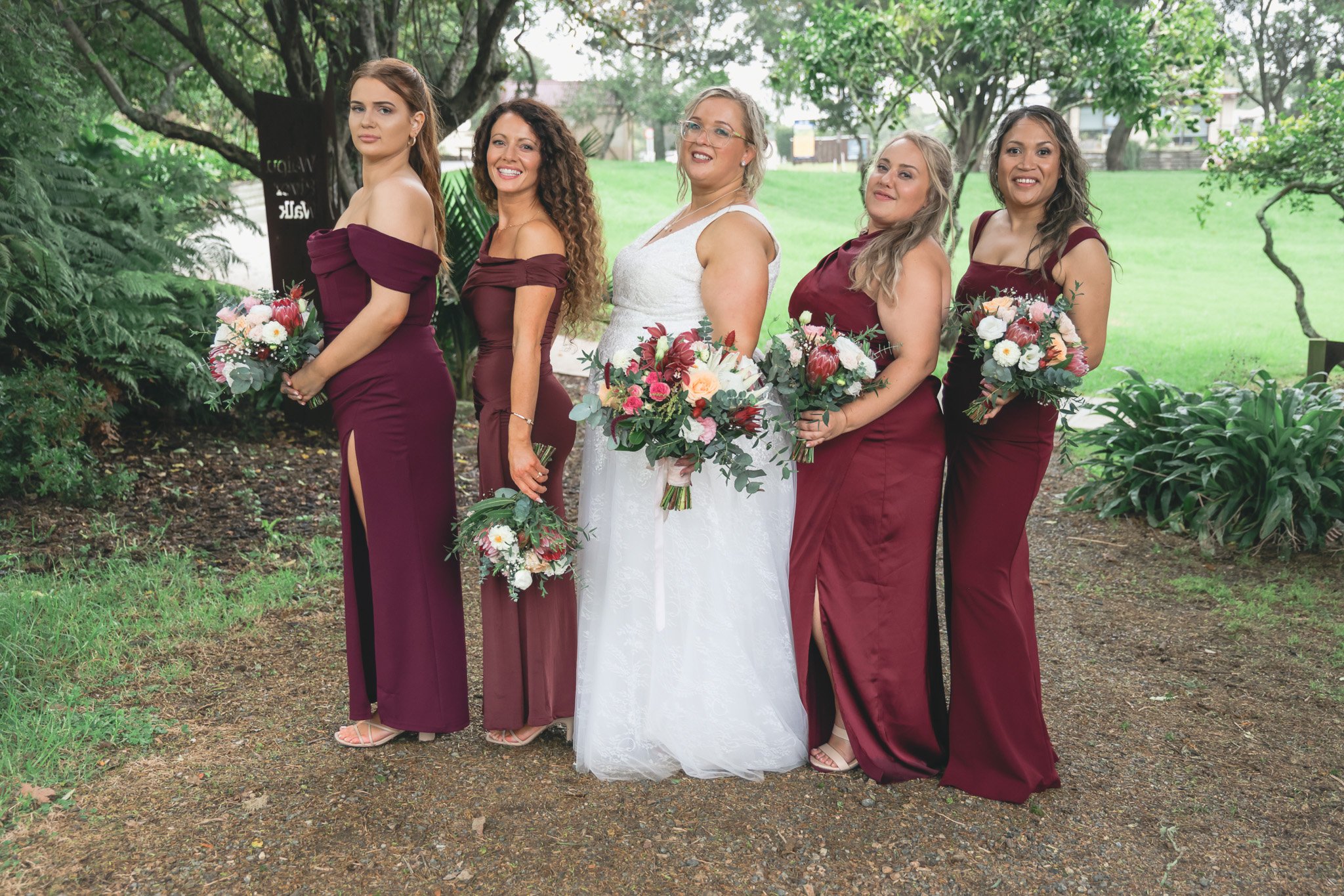 Group of six women in formal dresses standing outdoors in a park, holding bouquets of flowers, with trees and grass in the background.