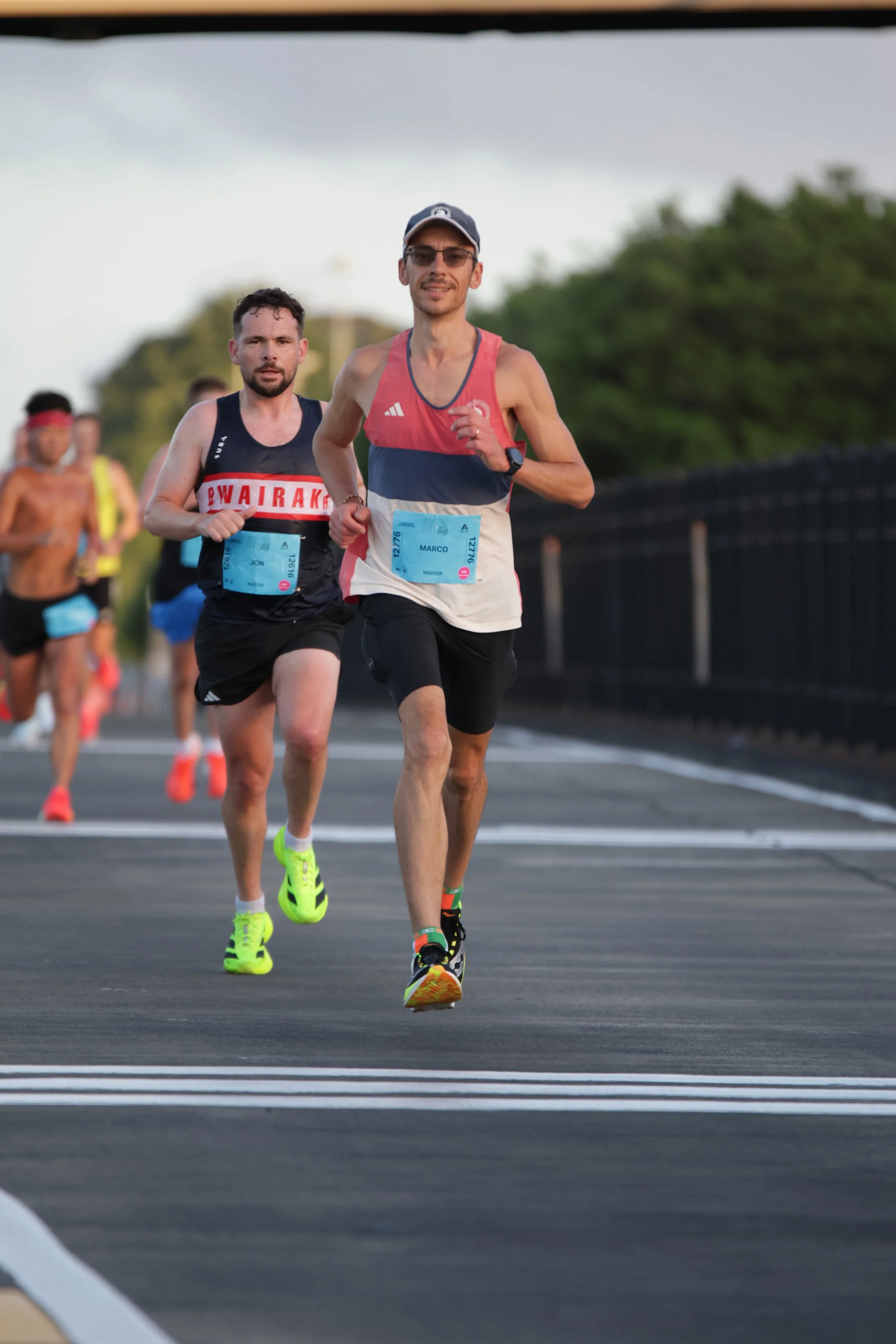 A group of runners participating in a race on a paved road, with trees in the background, during daytime. The leading runner is wearing a pink and white tank top, black shorts, and a cap. Behind him are other runners, including a shirtless man with a