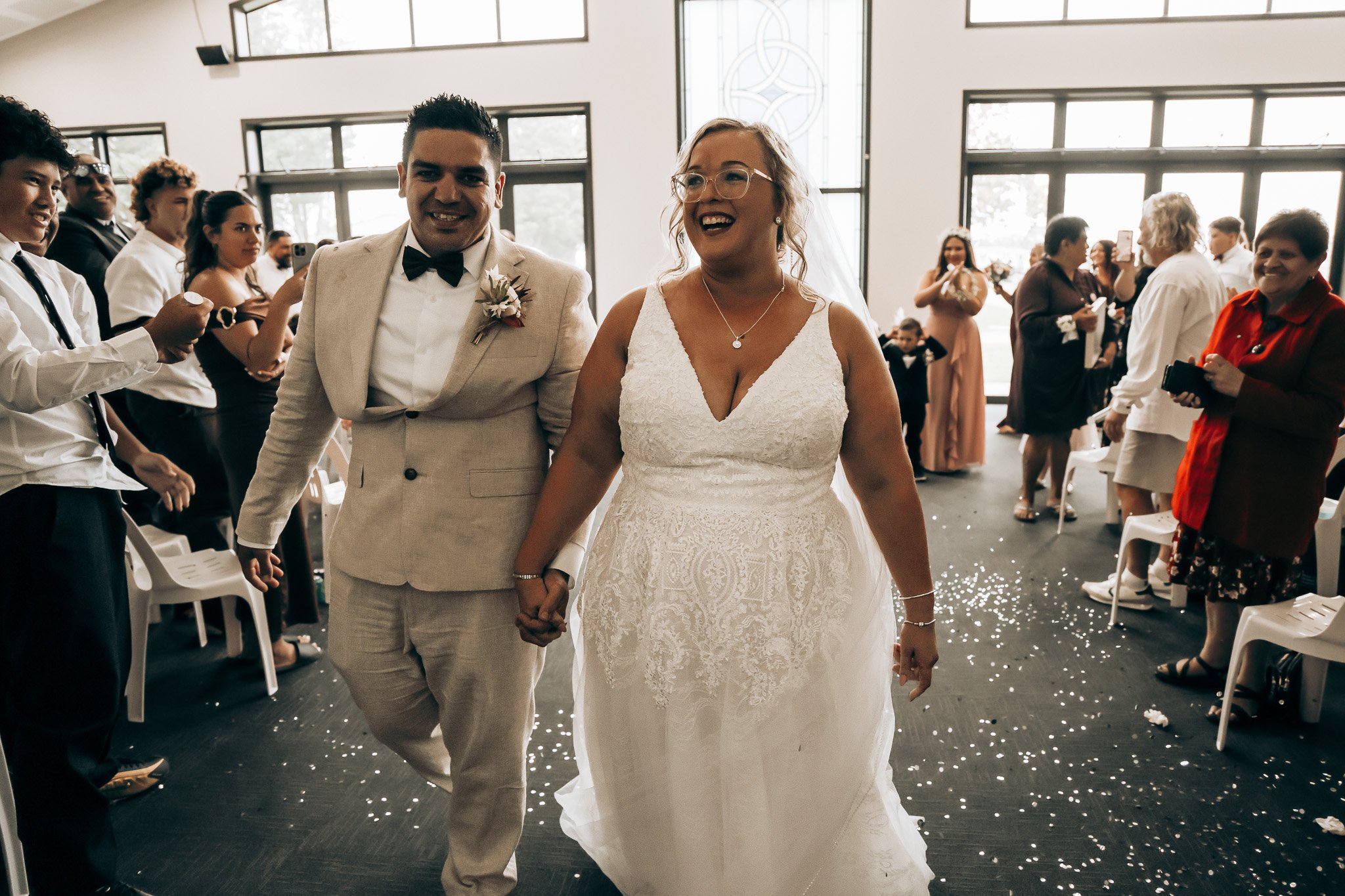 A bride and groom holding hands and smiling as they walk down the aisle at their wedding ceremony, surrounded by wedding guests in a well-lit indoor space.