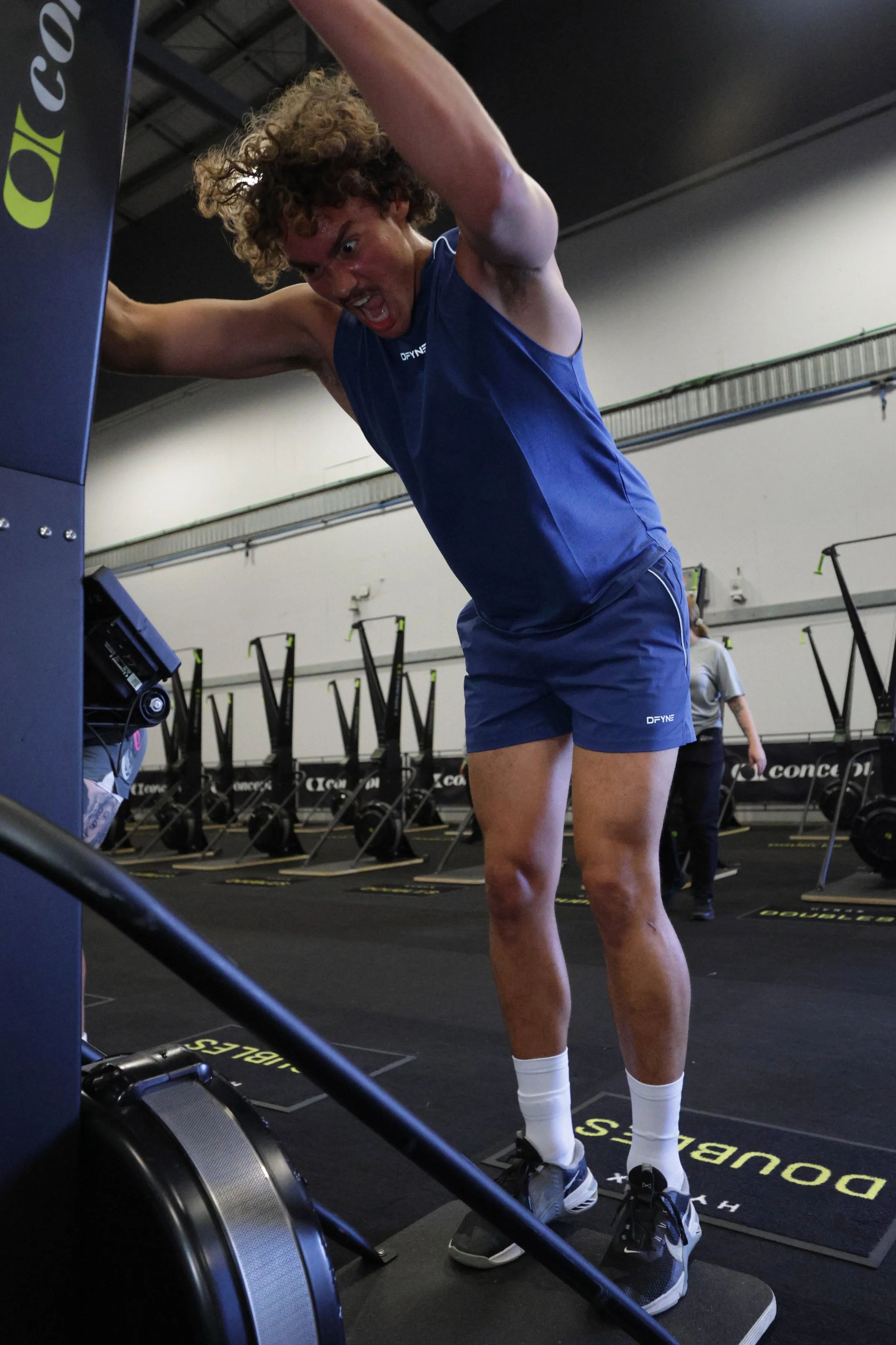 Man with curly hair wearing a blue sleeveless shirt and shorts, standing on a black workout mat with yellow markings, pushing a sled in a gym with rowing machines and other exercise equipment in the background.